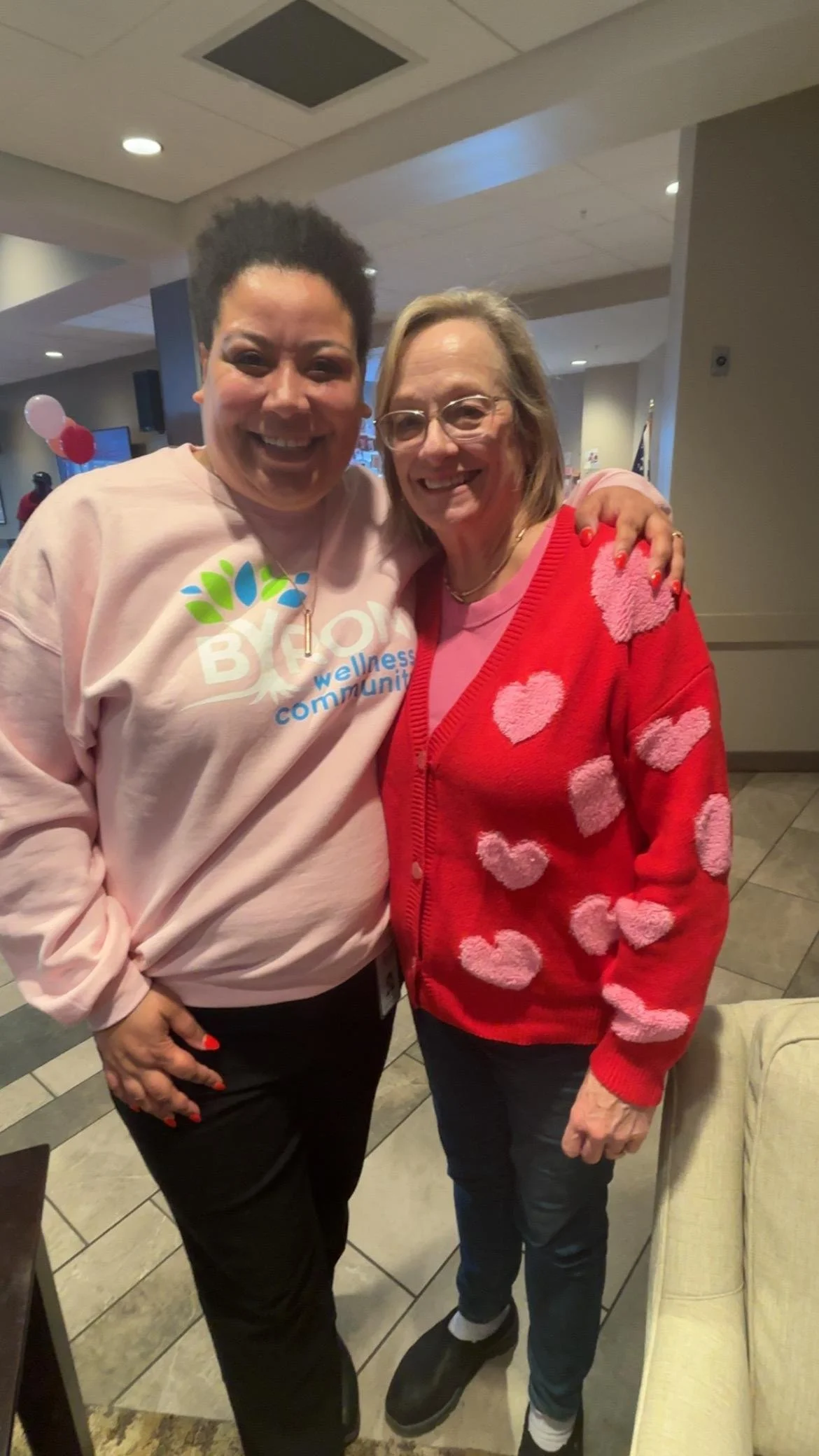 Two women smiling and posing together indoors. One woman has short curly black hair, wearing a pink sweatshirt with colorful print. The other woman has shoulder-length blonde hair, glasses, and is wearing a red sweater with pink hearts and a pink shirt underneath.
