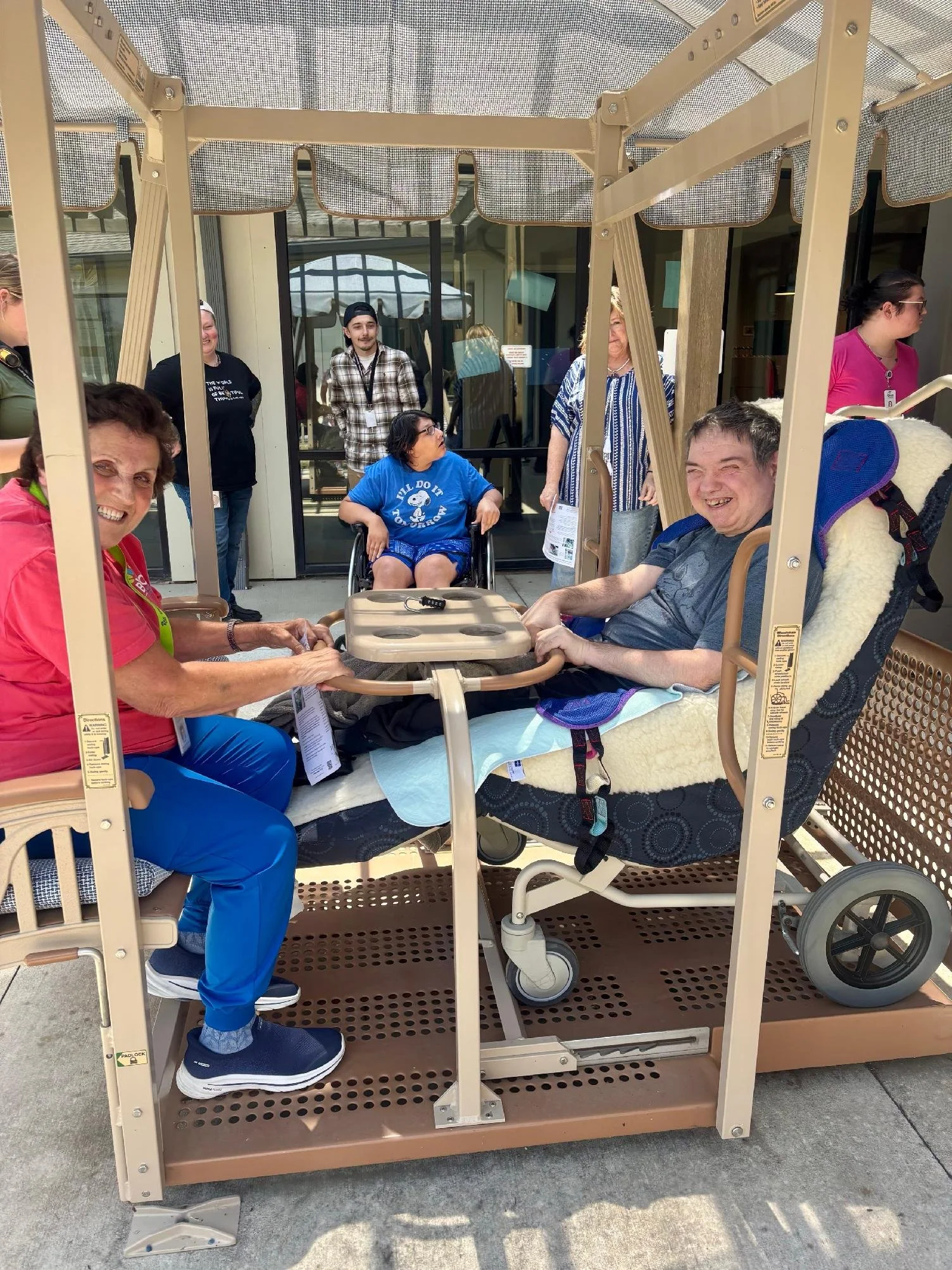 A group of people gathered around a wheelchair-accessible outdoor medical transport cart with a roof, situated outside a building with glass doors. Some are seated in the wheelchair, while others stand nearby and chat.