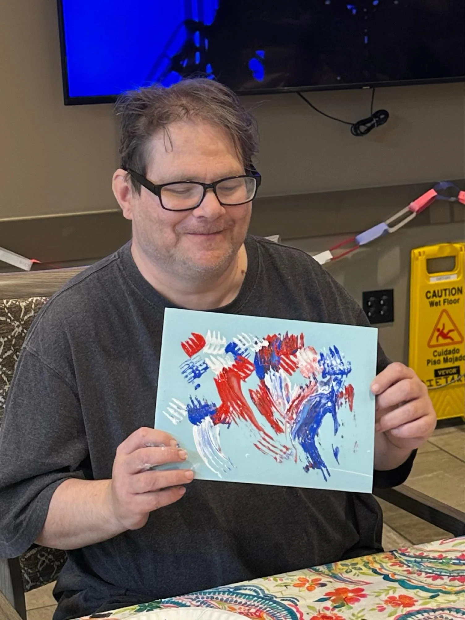 A man with glasses and unkempt hair is smiling and holding a colorful abstract painting that features red, white, and blue brushstrokes. He is sitting at a table covered with a floral tablecloth, with a yellow caution sign and a black electrical outl
