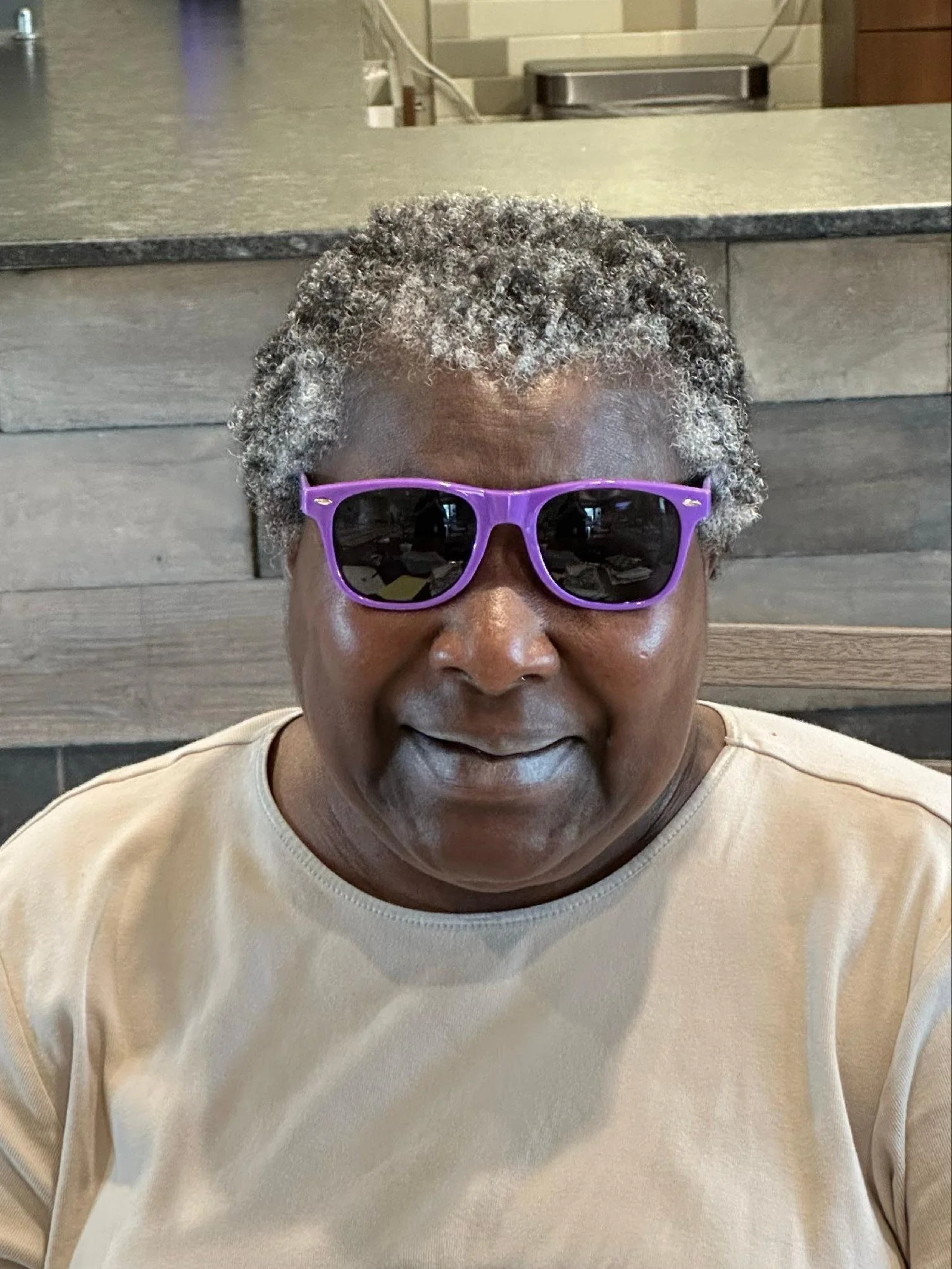 Close-up of an elderly person with curly gray hair and dark sunglasses, wearing a beige shirt, sitting indoors against a wooden wall.
