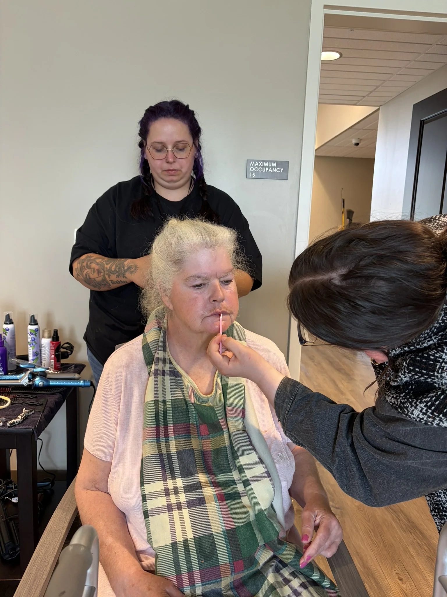 An elderly woman with blonde hair sitting in a brown chair, having her face makeup applied by a makeup artist. The woman wears a pink shirt with a green and pink plaid scarf. The artist is using a pink brush near her lips. There are various makeup su