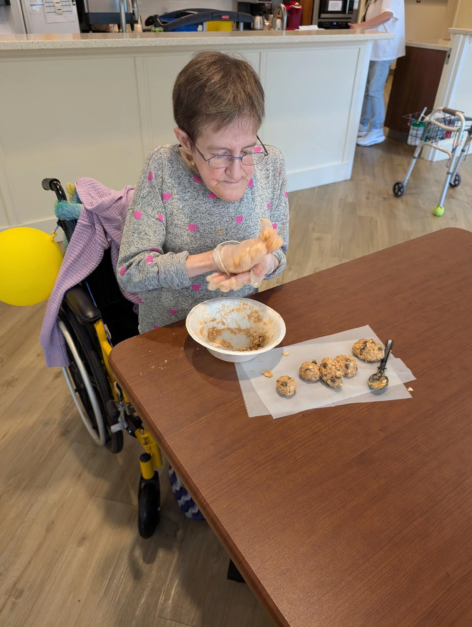 An elderly woman in a wheelchair sits at a table, handling baked cookies on a paper-lined tray. In the background, a person stands at a kitchen counter, and a walker with a tennis ball on one leg is visible.