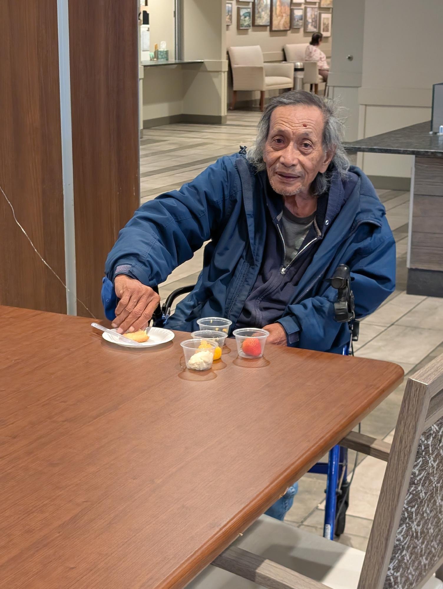 An elderly man with gray hair, wearing a blue jacket, sits at a wooden table with small cups of assorted desserts, in what appears to be a cafe or dining area. In the background, a woman in pink sits at another table, with framed pictures on the wall