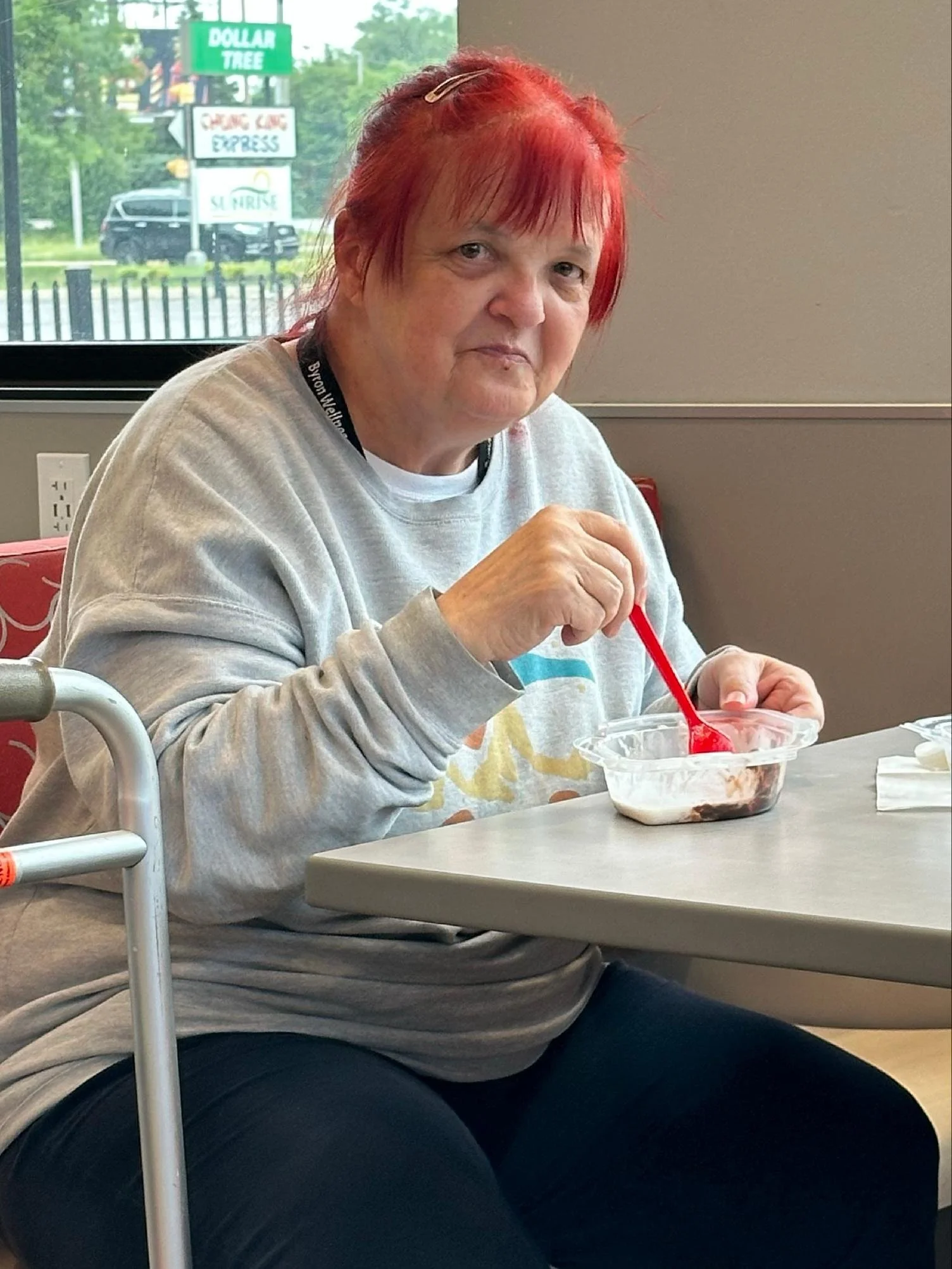 A woman with red hair and a hair clip sitting at a table in a restaurant or cafe, holding a red spoon and eating dessert from a clear plastic bowl.