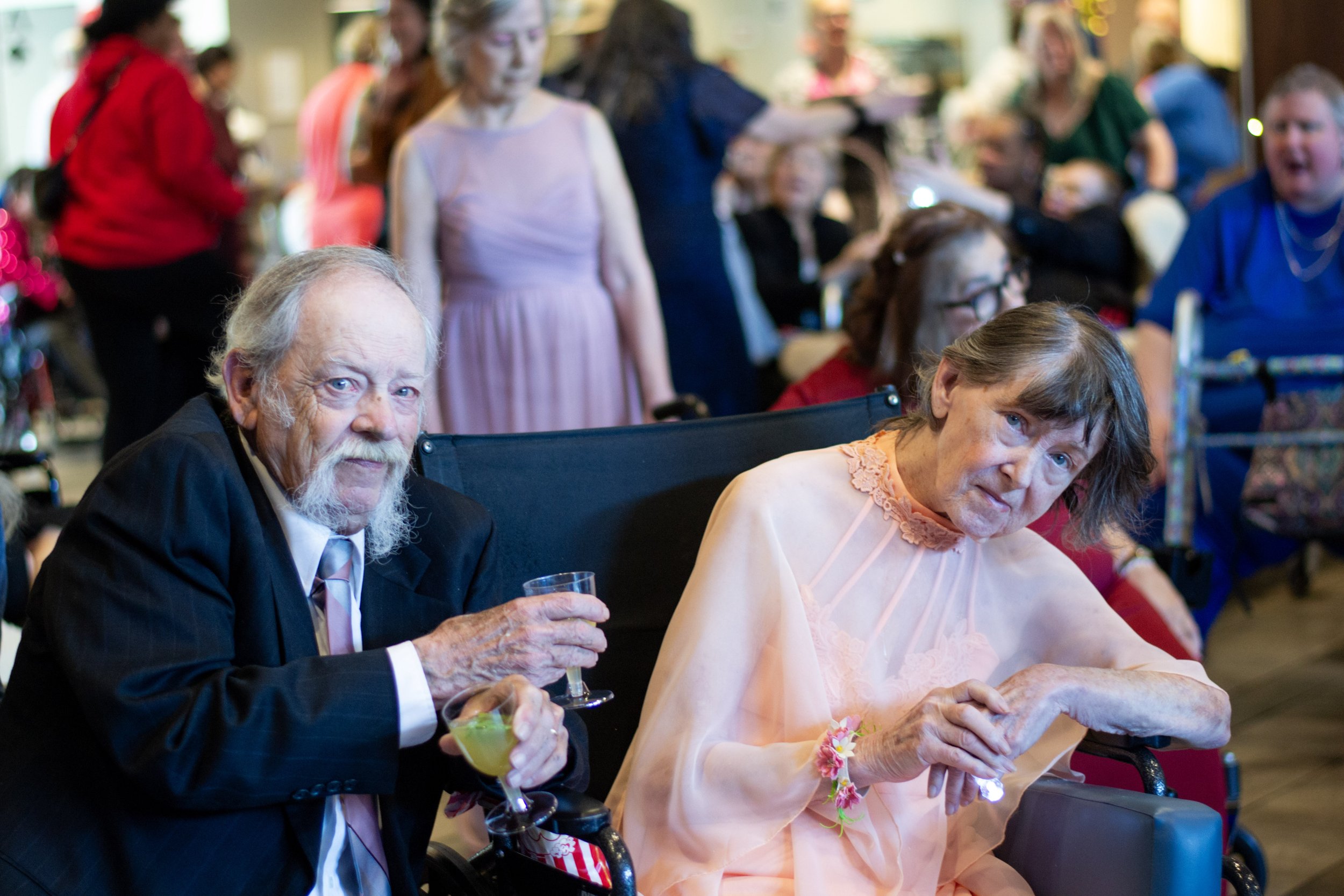 Older man with white hair and beard in a suit holding a drink, sitting next to an older woman in a peach dress with floral details, both at a social gathering.