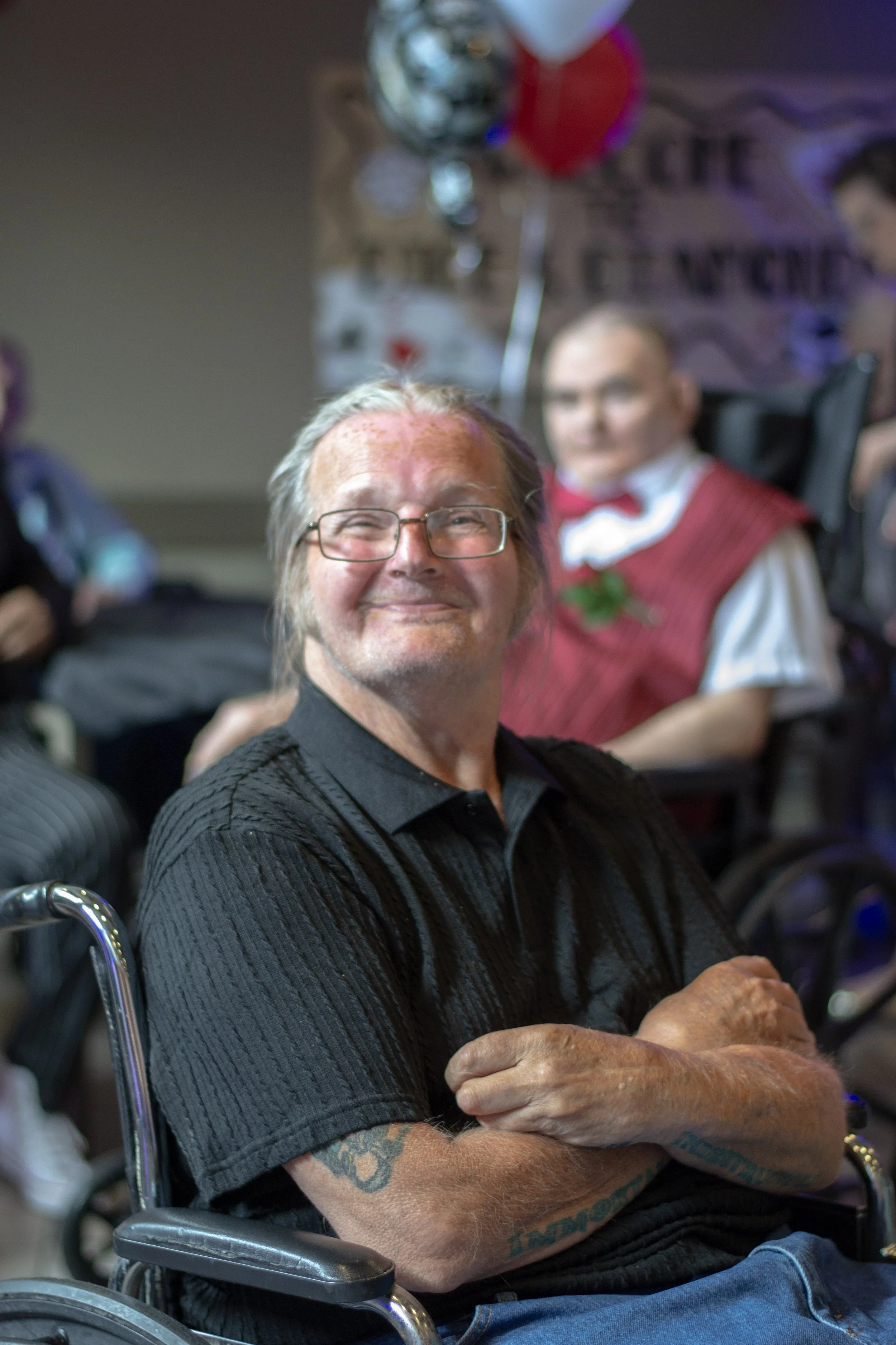 A smiling man with glasses and tattoos on his arms, sitting in a wheelchair with his arms crossed, in a room with balloons and party decorations, including a person dressed in a red and white vest with a boutonniere in the background.