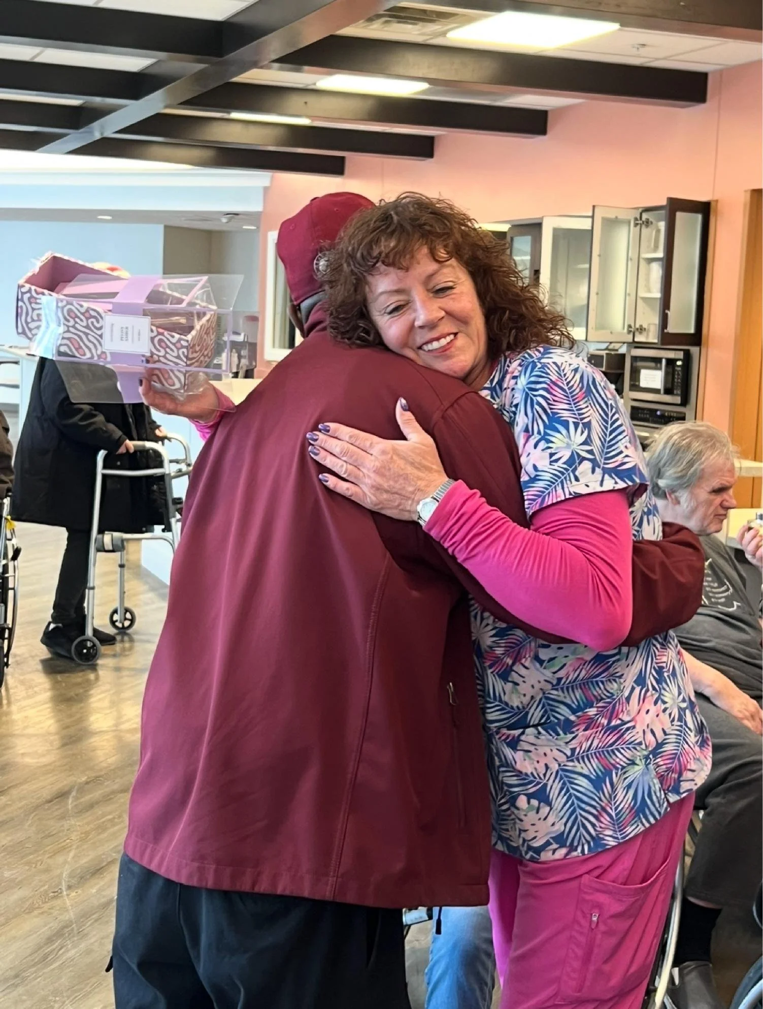 A woman with curly hair smiling and hugging a person wearing a red jacket in a room with people in the background, including a person with a walker and another with gray hair sitting in chairs.