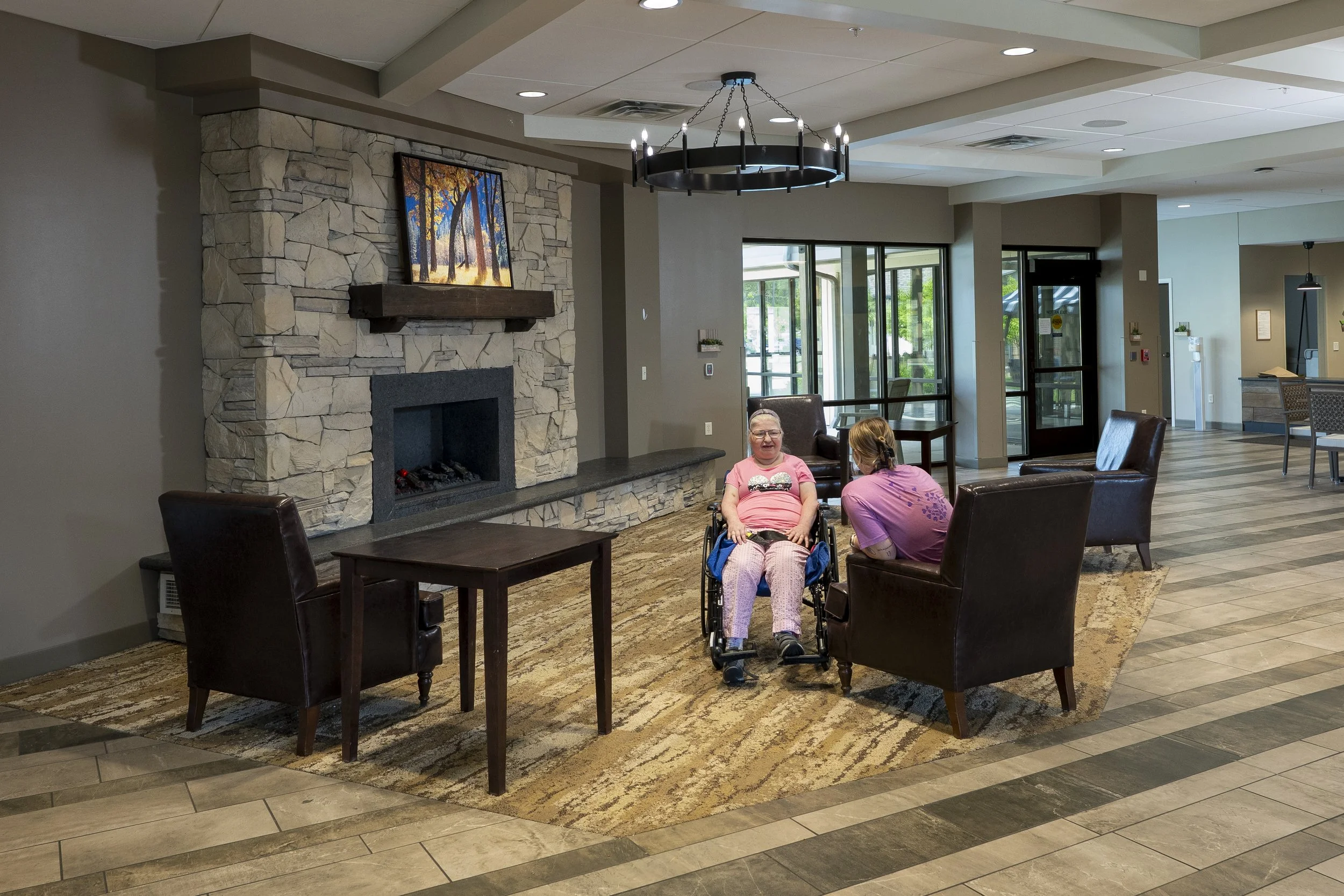 A spacious, modern lobby with a stone fireplace, wall-mounted TV, and comfortable chairs. Two women, one in a wheelchair, are sitting and talking. Large windows allow natural light, and there is a chandelier hanging from the ceiling.