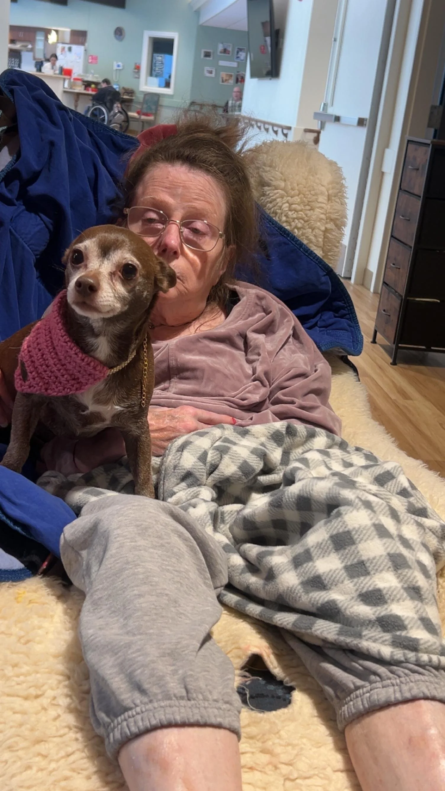 An elderly woman with glasses lying on a couch with a small brown dog wearing a pink sweater in her arms. The woman and dog are on a cream-colored shaggy blanket in a cozy room.