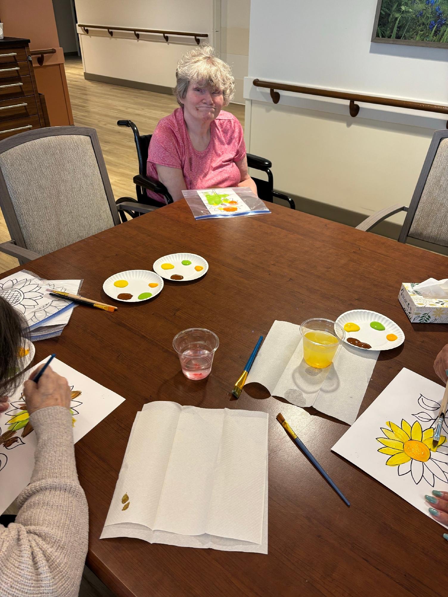 An elderly woman with curly gray hair wearing a pink shirt, sitting in a wheelchair, participating in an arts and crafts activity at a wooden table. The table has paper with sunflower coloring pages, paint palettes, brushes, and cups of water and jui