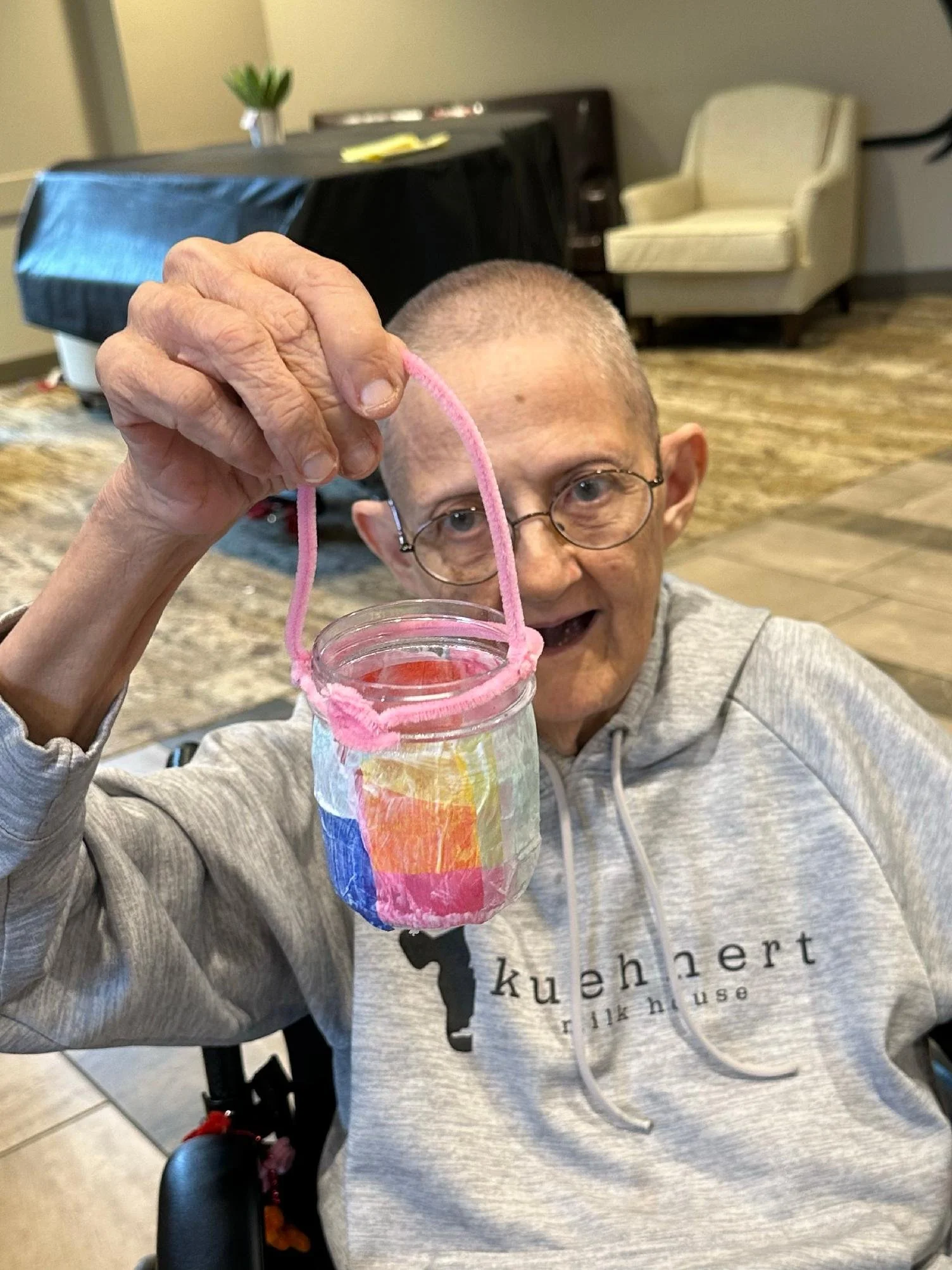 An elderly person with glasses holding a colorful craft made from a plastic bottle and pink pipe cleaner.