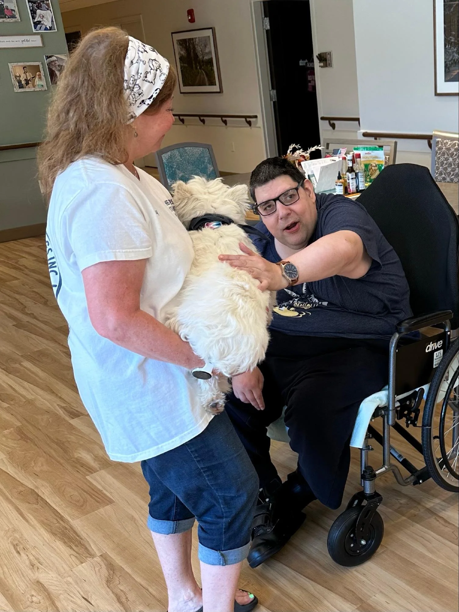A woman and a man with a service dog in a wheelchair greeting each other indoors.