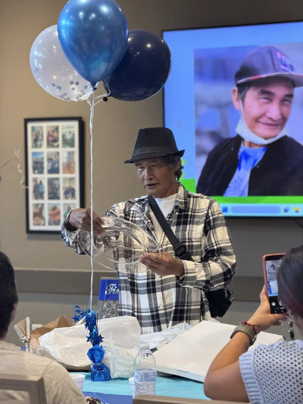An elderly woman wearing a black hat and plaid shirt opening a clear plastic gift, surrounded by balloons and a group of people, with a large screen displaying a smiling young man in the background.
