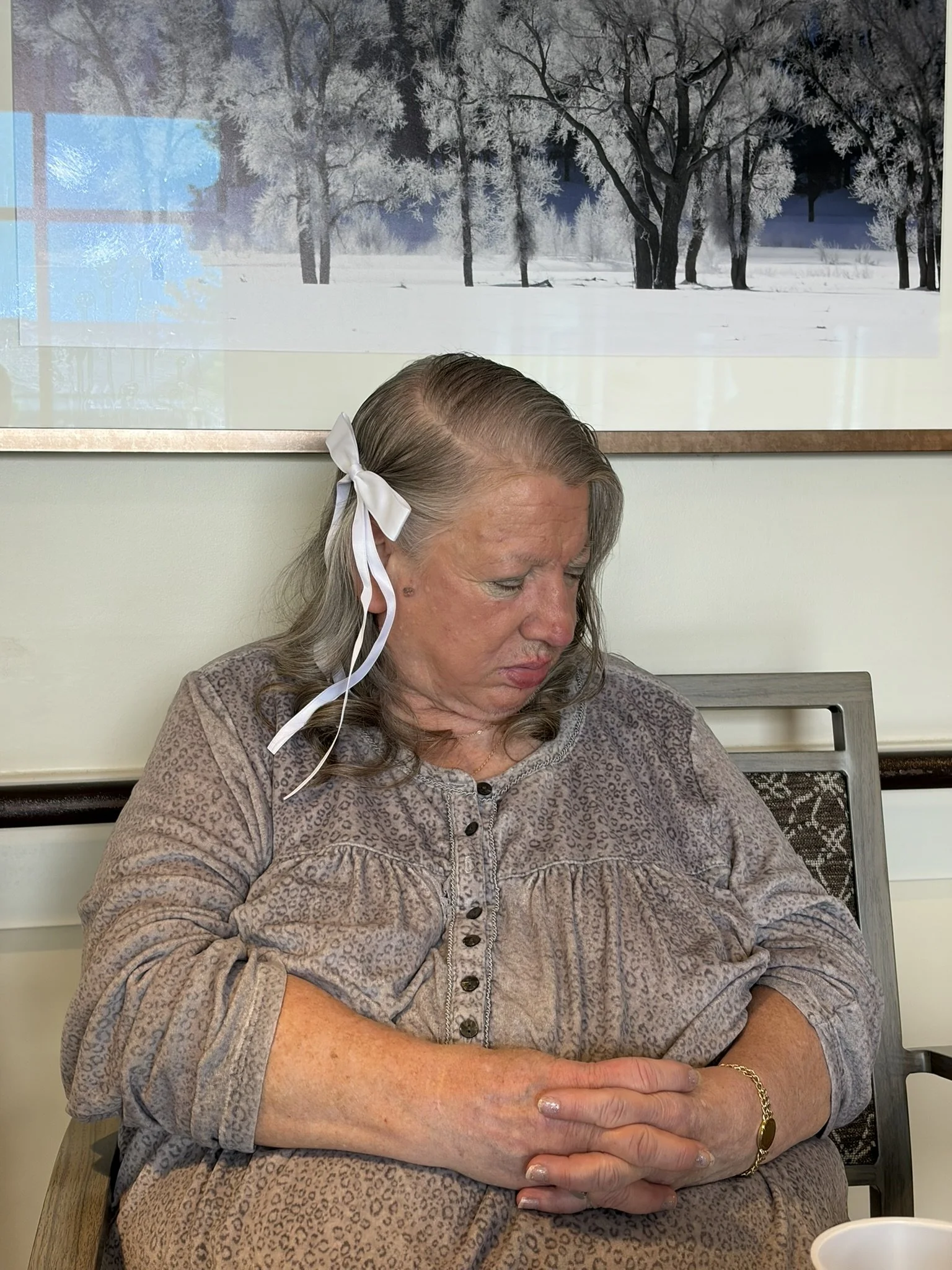 An elderly woman with long gray hair tied with a white ribbon, sitting at a table with her arms crossed, wearing a leopard print shirt and a gold bracelet. Behind her, a large framed picture of a snow-covered landscape with trees is hanging on the wa