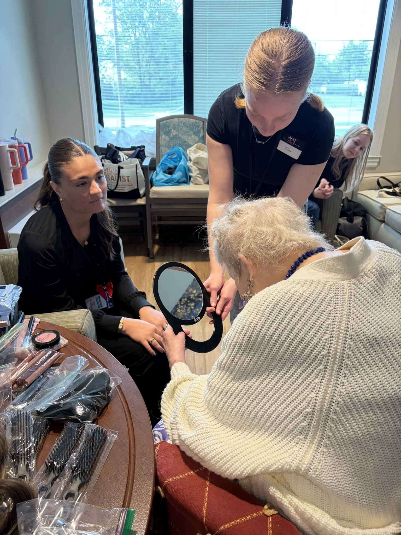 A woman with light brown hair styled in pigtails appears to be performing a manicure or nail treatment on an elderly woman with blonde hair. Two other women are observing in the background. The scene takes place in a bright room with large windows, a
