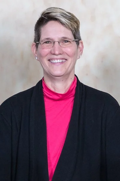 A woman with short hair wearing glasses, a pink shirt, and a black cardigan, smiling in front of a neutral background.