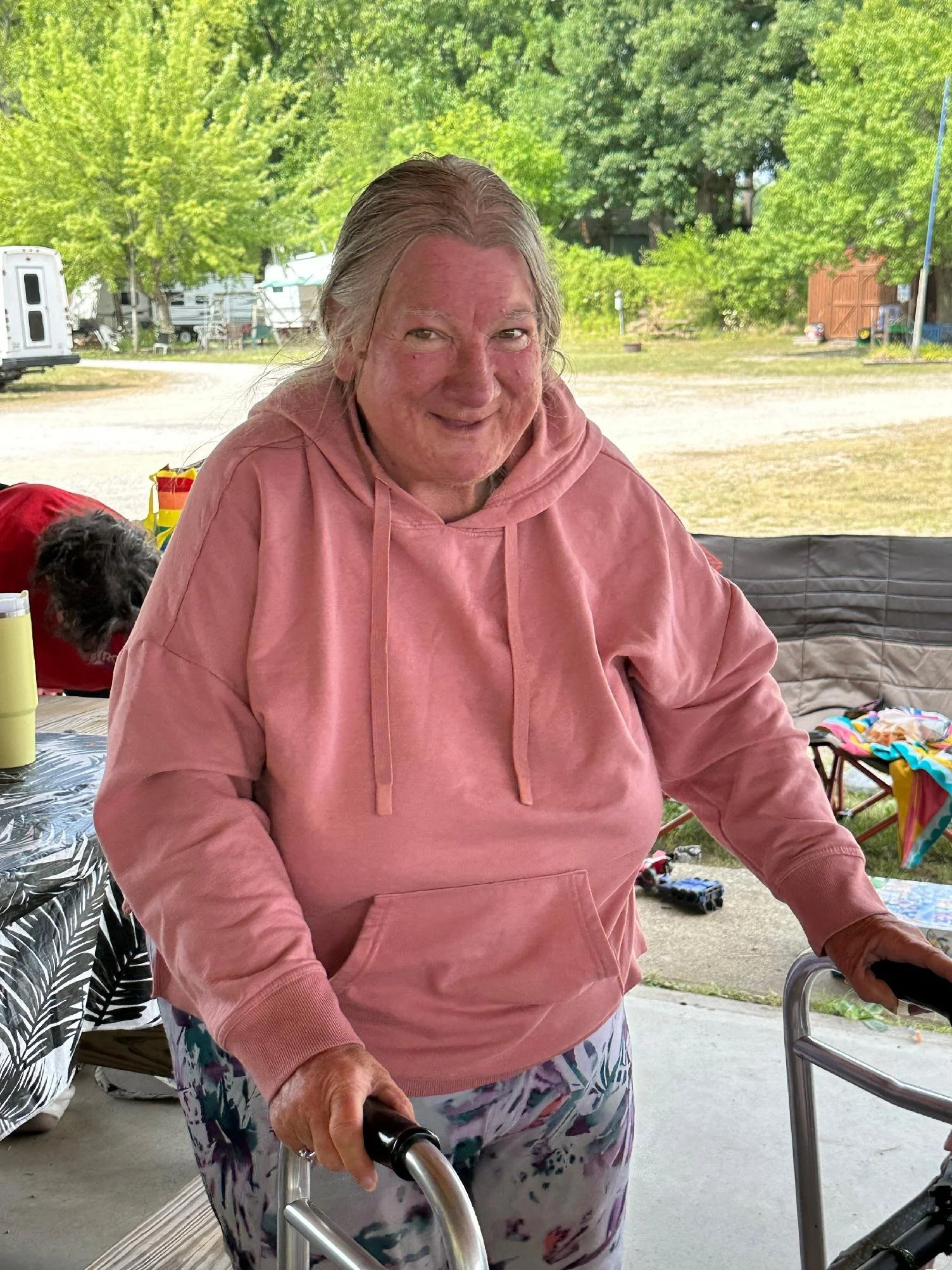 An elderly woman with long gray hair, wearing a pink hoodie and colorful pants, standing outdoors with a walker. There is a smiling person with long gray hair and a dog in the background, along with trees, a camper, and outdoor chairs.