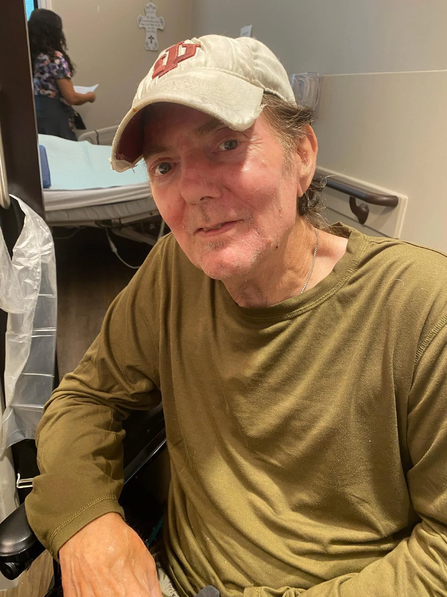 A middle-aged man wearing a beige baseball cap with a red N on it, and a green long-sleeve shirt, sitting in a medical setting with a hospital bed and a woman in the background.