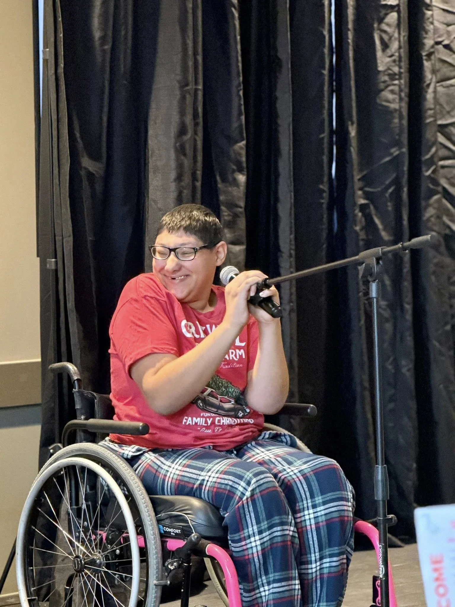 A young man with glasses smiling and holding a microphone, sitting in a wheelchair with a plaid shirt and patterned pants, in front of black curtains.