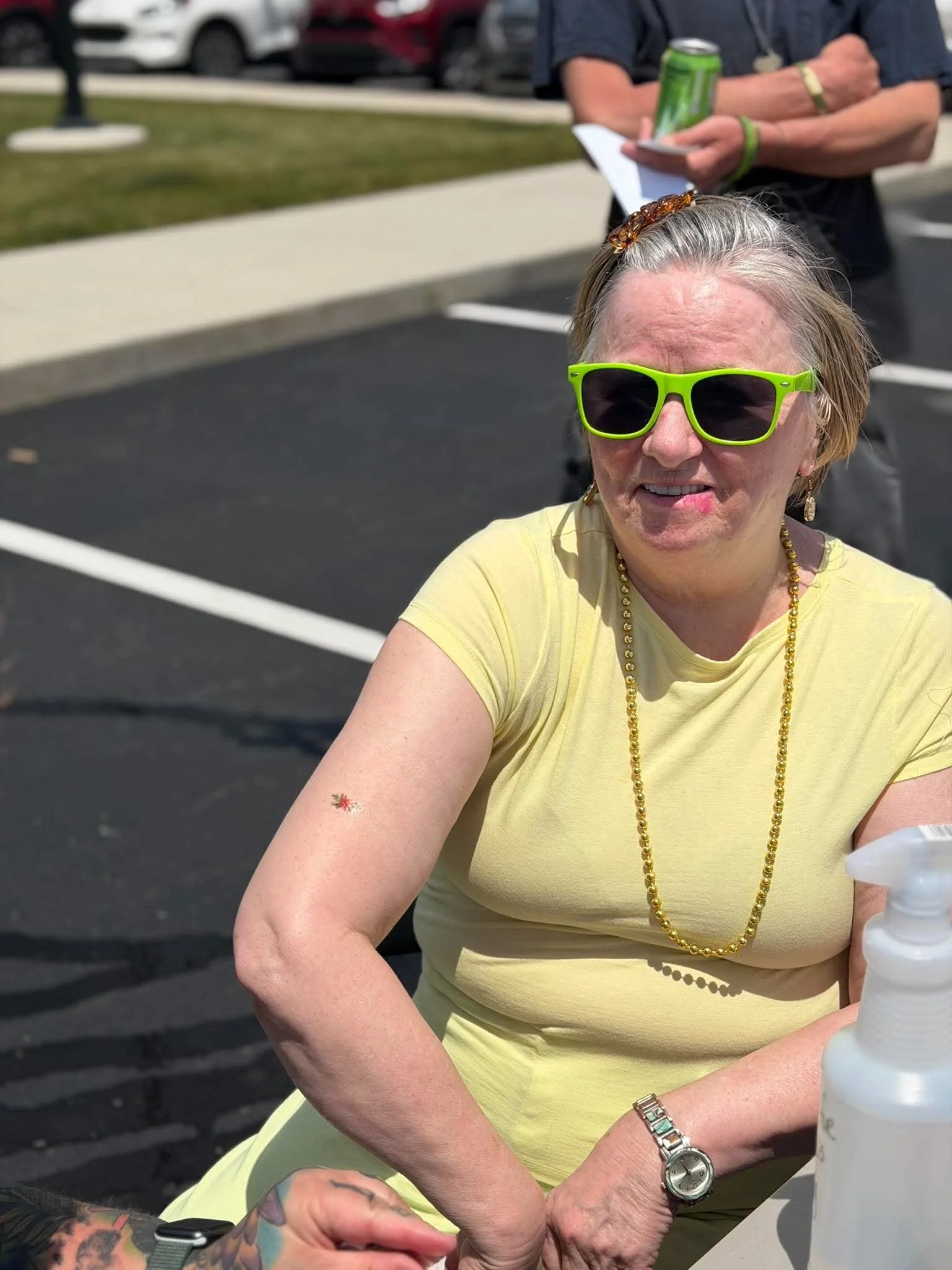 An elderly woman with gray hair, wearing bright green sunglasses, a yellow shirt, gold jewelry, and a watch, sitting outdoors at a table with a small band-aid on her arm, with a person standing behind her holding a can and a phone, in a parking lot.