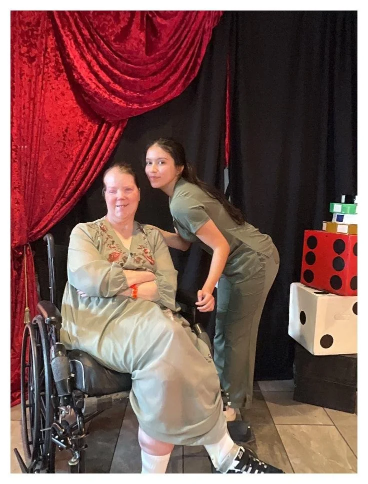 A woman in a wheelchair and a young girl in scrubs posing together in front of a red and black curtain, with large decorated blocks resembling dice on the right.