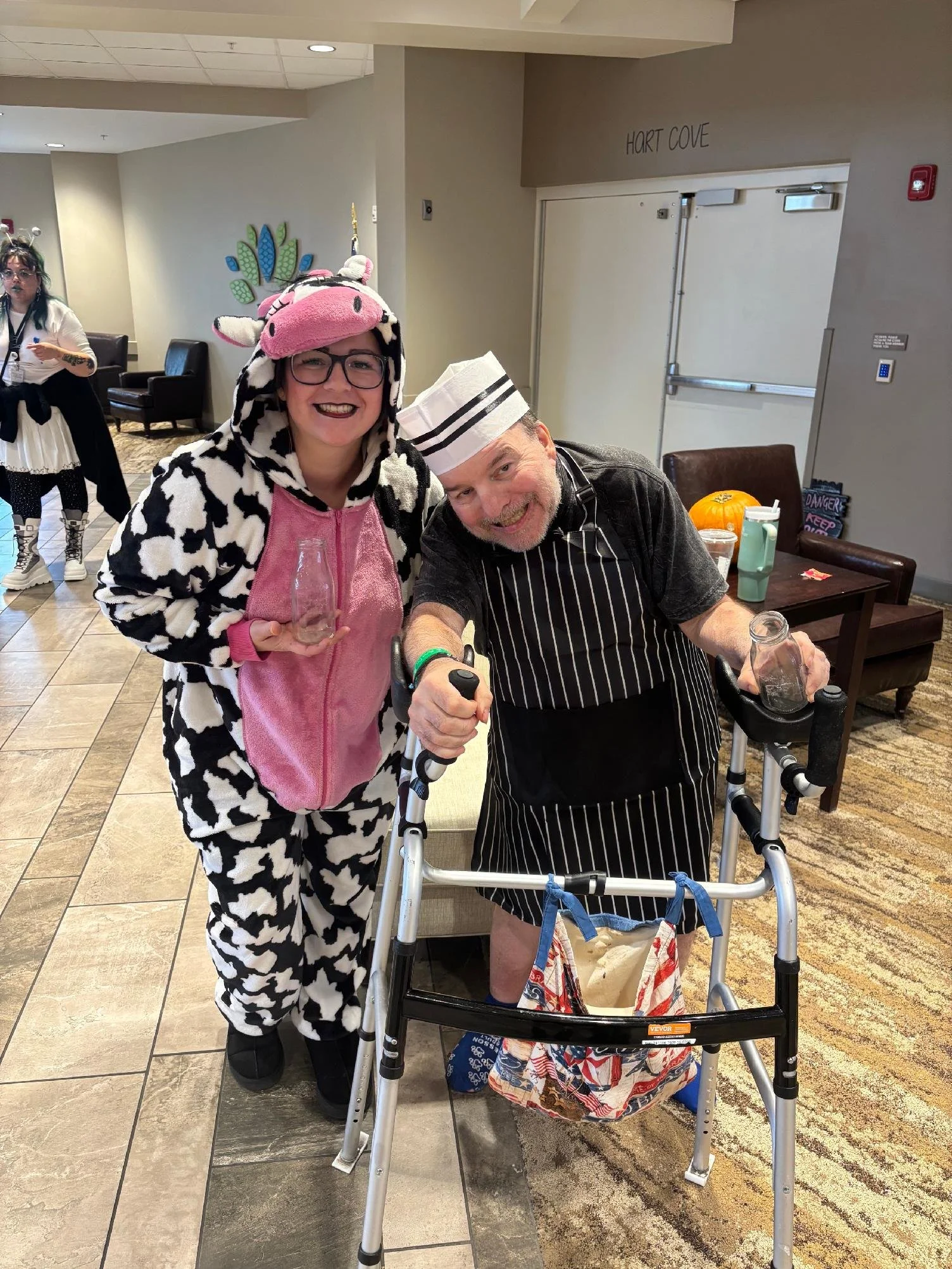 A woman in a cow costume and a man in a chef's outfit are smiling at the camera, with the woman holding a small glass bottle. The man is using a walker. They appear to be in a hospital or care facility lobby decorated with seasonally themed items like a pumpkin, and there are other people in costumes in the background.