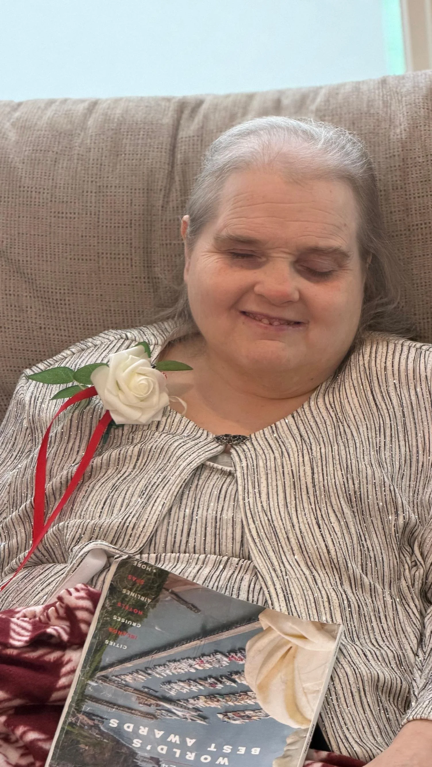 An elderly woman with gray hair sitting on a beige couch, wearing a striped outfit with a white flower and red ribbon pinned to it. She has her eyes closed and a gentle smile on her face. There is a magazine in her lap.