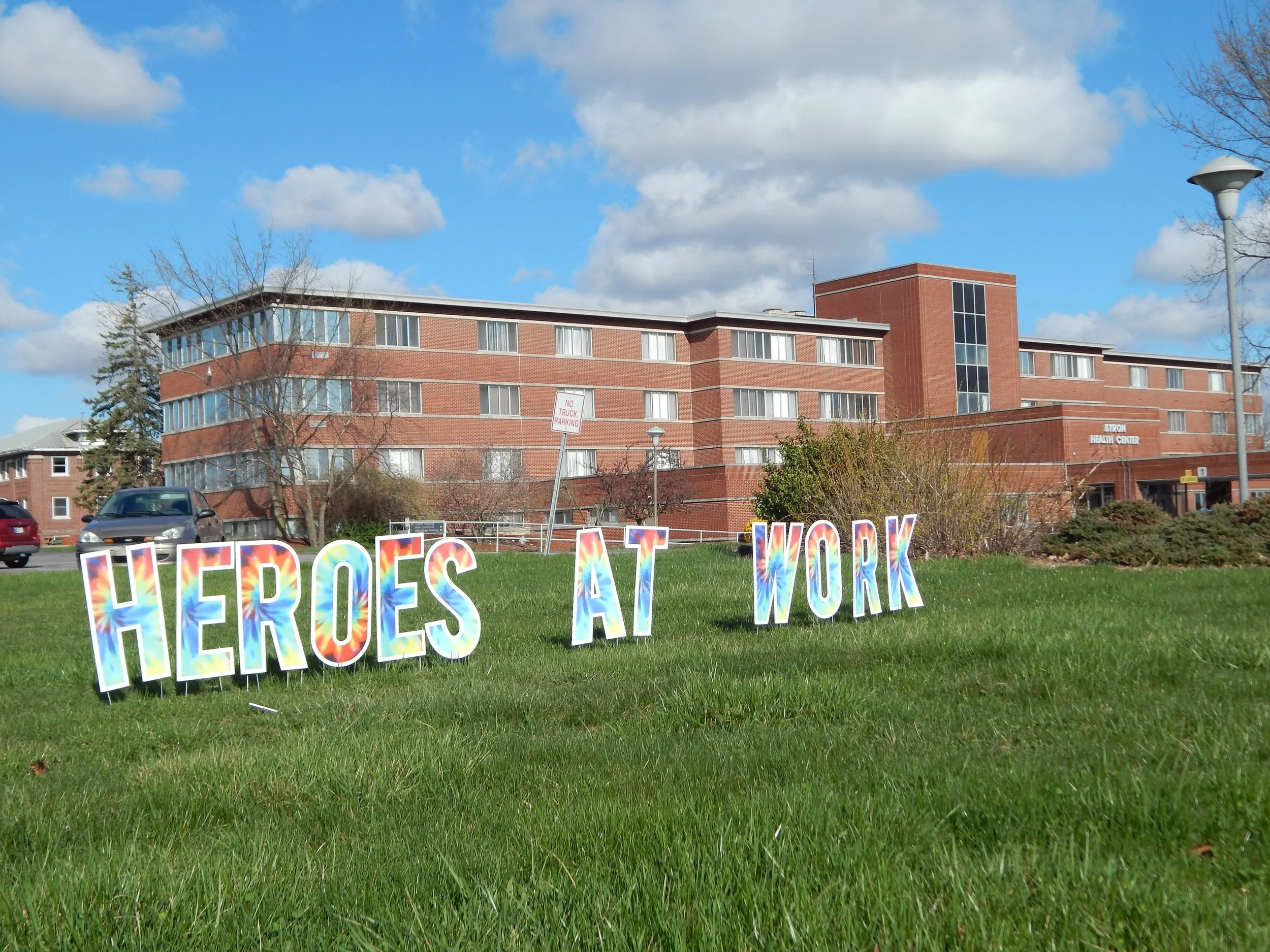 Colorful letters spelling out "HEROES AT WORK" on a grassy field outside a brick building with large windows and a sign for Byron Health Center, under a partly cloudy sky.