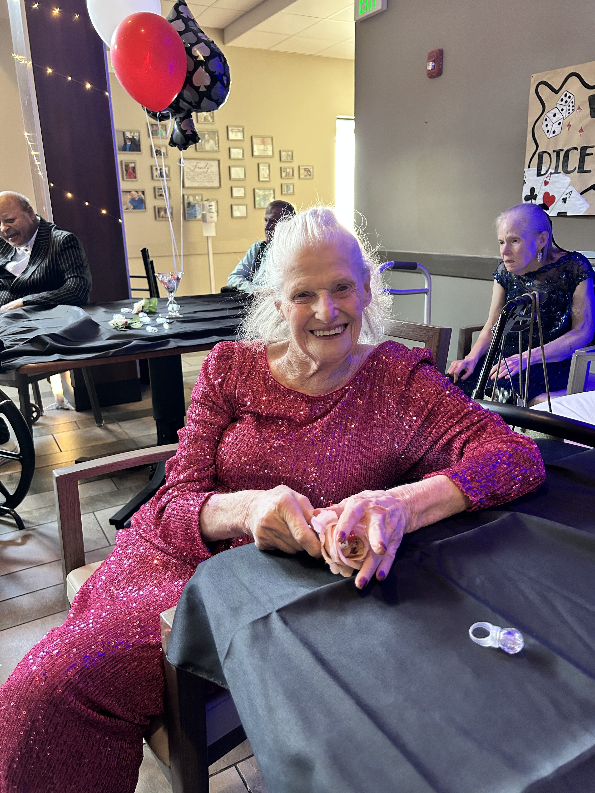 An elderly woman in a sparkly red dress smiling at a table during a celebration, with balloons and decorations in the background.