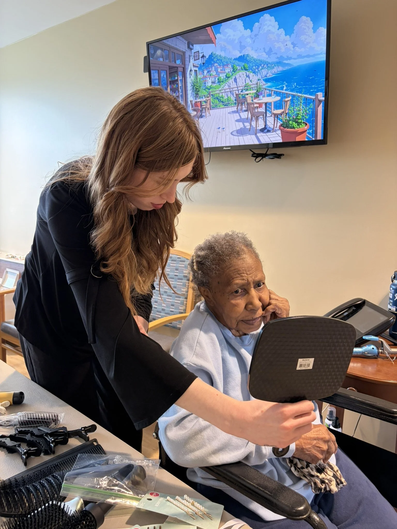 A woman with long hair assisting an elderly woman with gray hair in a wheelchair during a haircut or grooming session. They are in a room with a television showing a coastal scene and various hair styling tools and supplies on a nearby table.