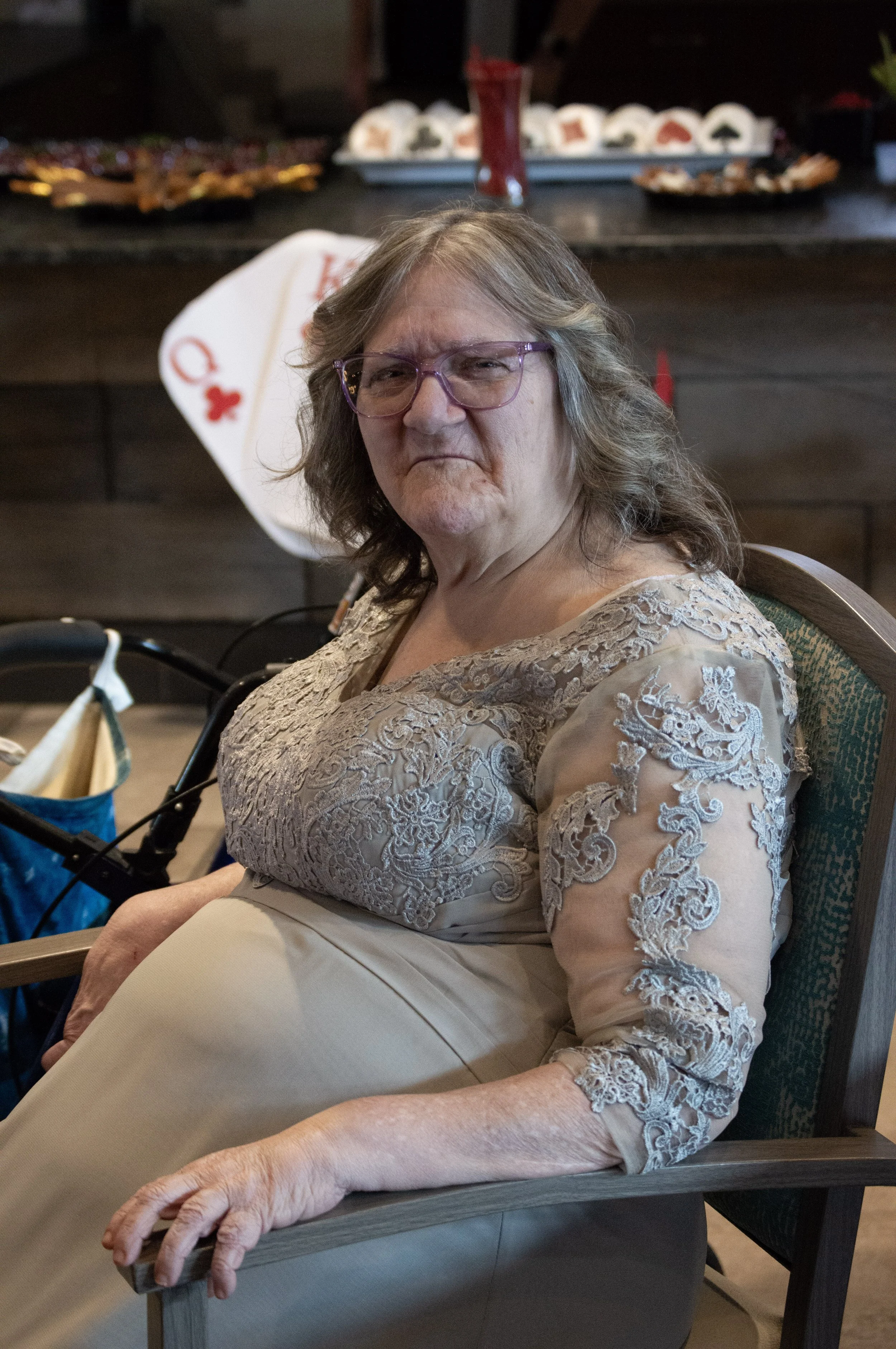 A senior woman with glasses and wavy gray hair seated at a table, wearing a lace dress, with food in the background and a gaming table decoration.
