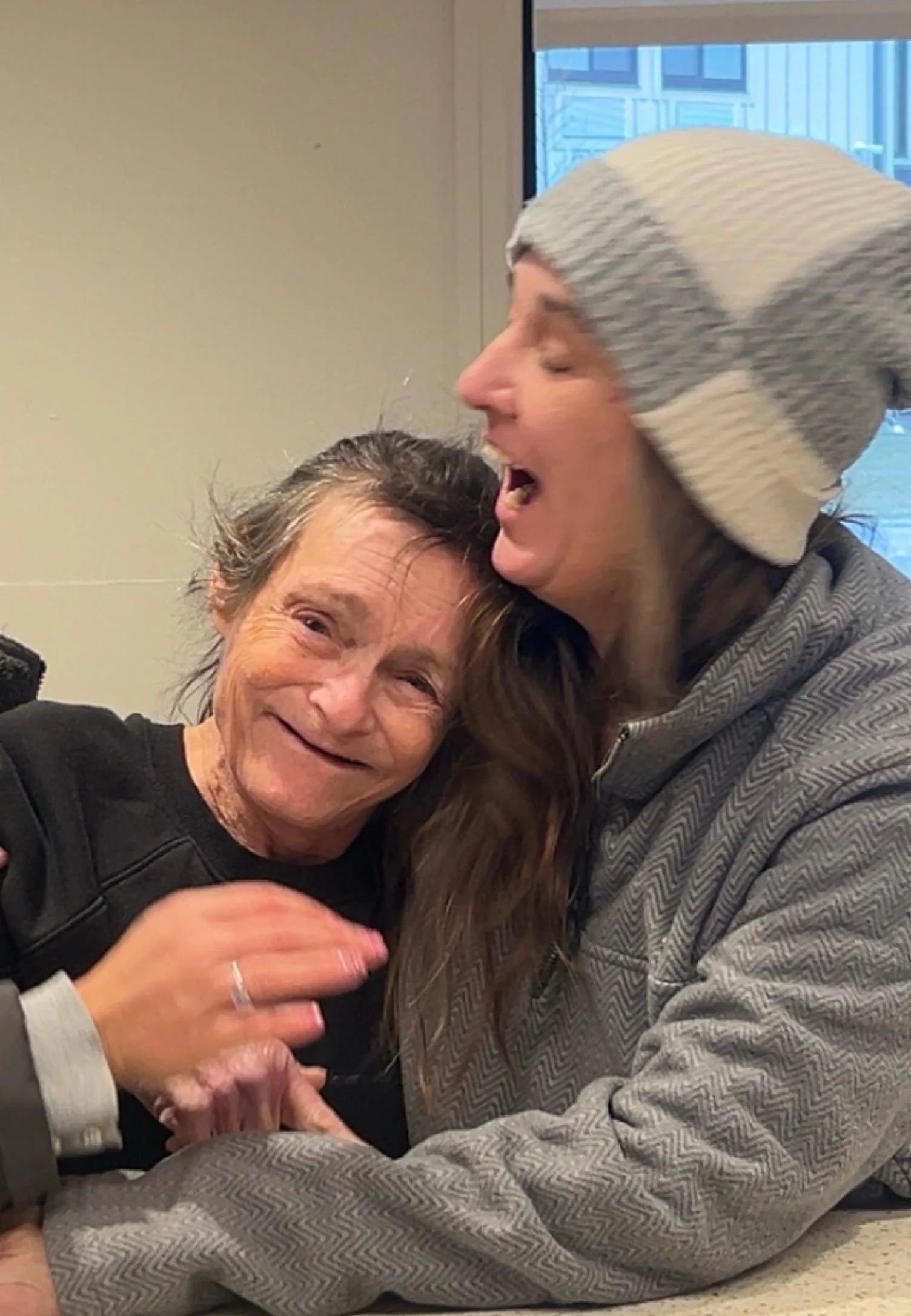 Two women sharing a joyful moment, one of them appears to be older, and the other younger, both smiling, with the younger woman wearing a gray and white knit cap and a matching jacket.