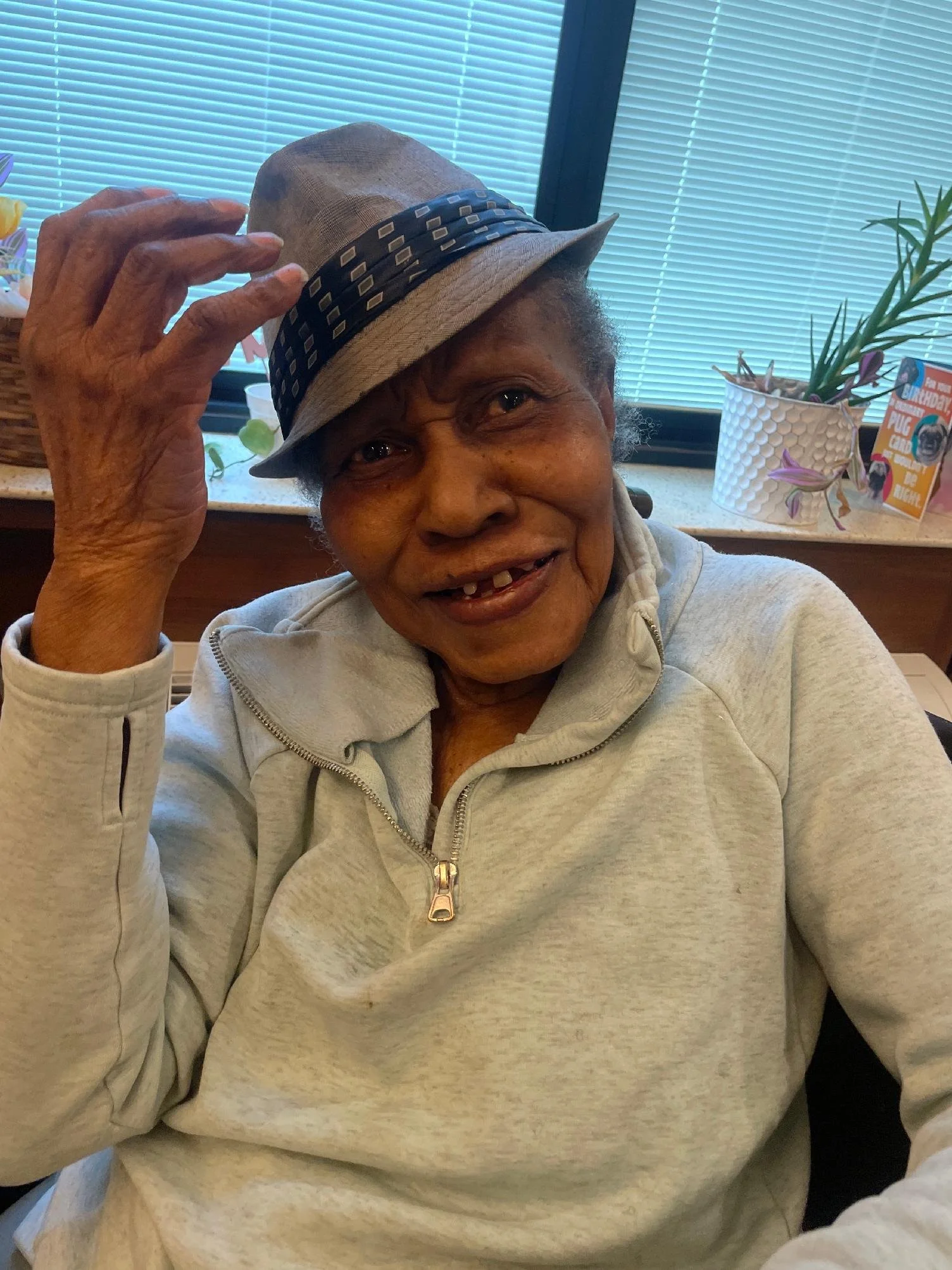 A smiling elderly woman with missing teeth, wearing a gray zip-up hoodie and a beige hat with black and white accents, sitting indoors near a window with blue blinds, with a plant and a birthday card in the background.