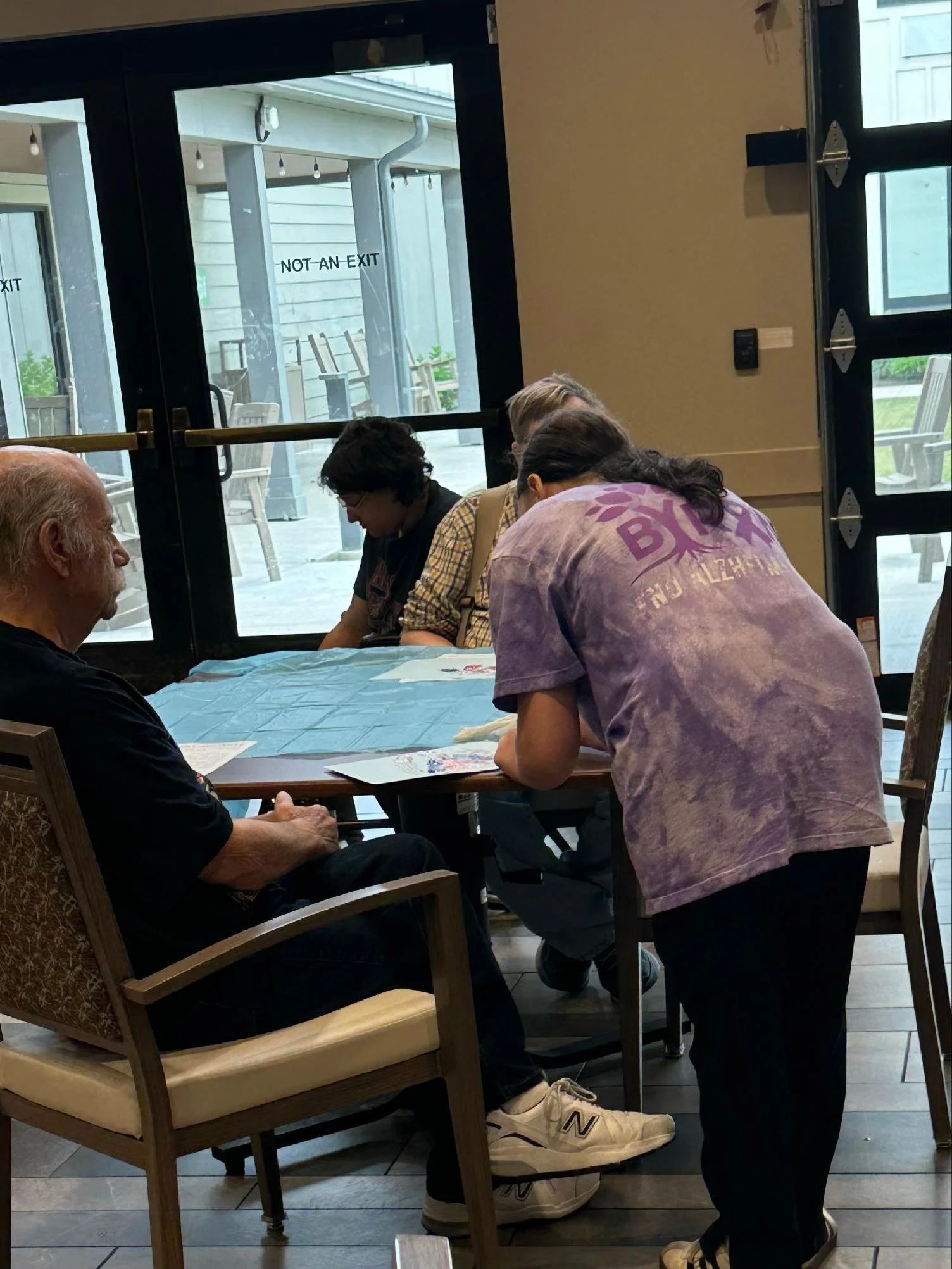 A group of people sitting around a table inside a restaurant, with a person in a pink shirt standing at the table. Through the window, there are outdoor chairs and a porch with a sign that says "NOT AN EXIT."