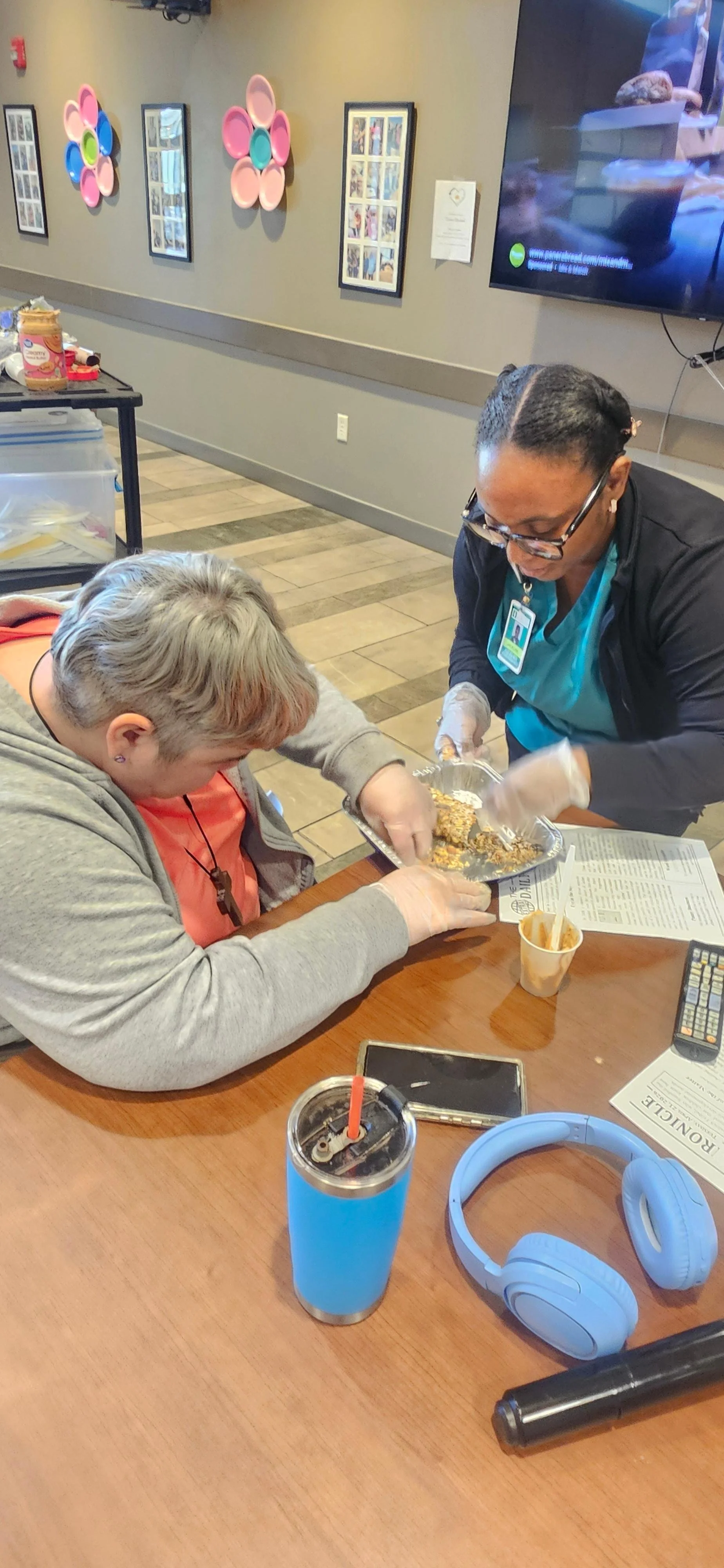 A woman serving food to a person at a table in a community center or cafe setting, with colorful wall decorations, a TV, and various personal items on the table, including a blue tumbler, headphones, and a smartphone.