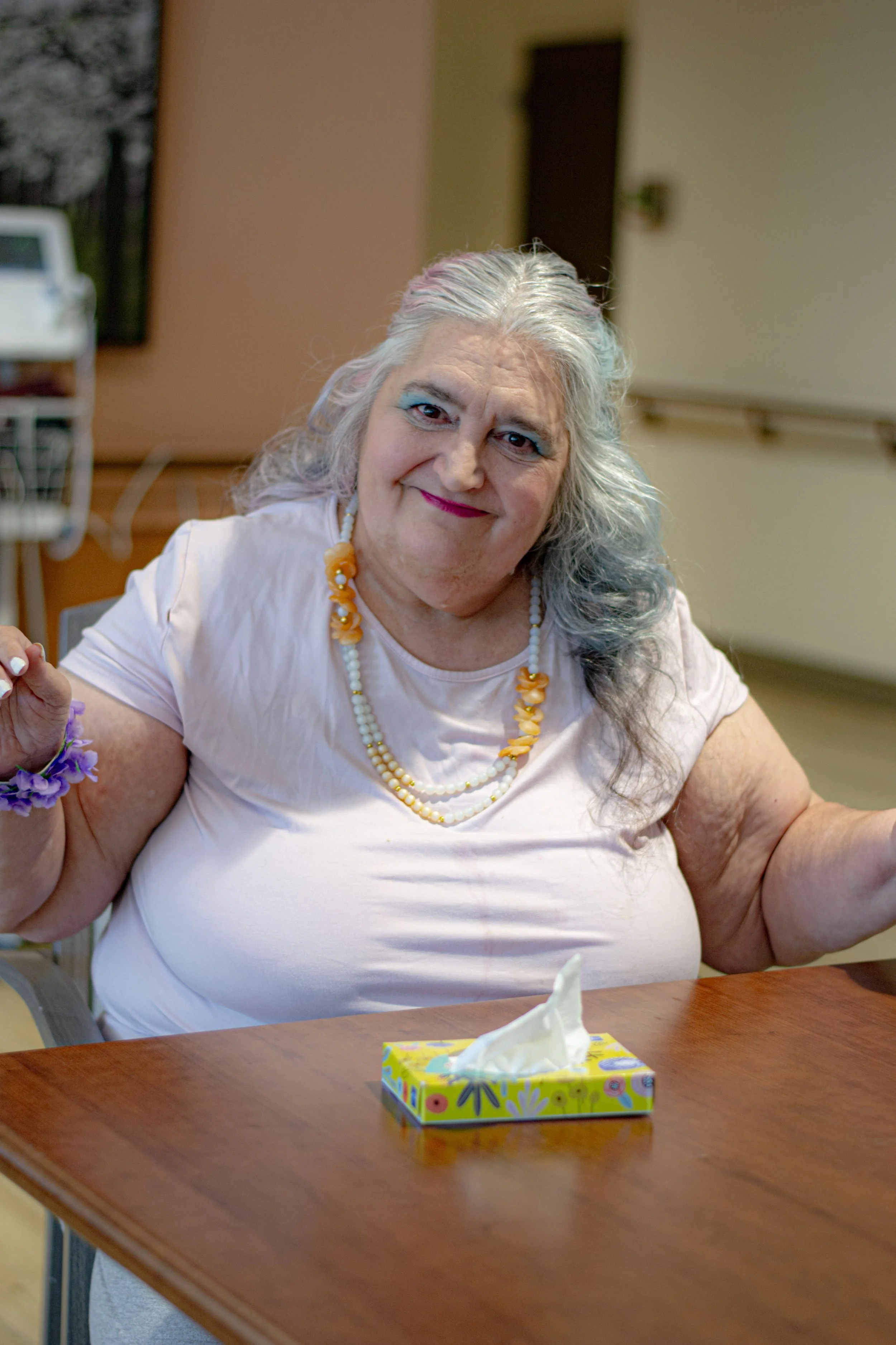 An elderly woman with gray hair, wearing a white shirt and yellow-bead necklace, sitting at a wooden table with a colorful tissue box in front of her.