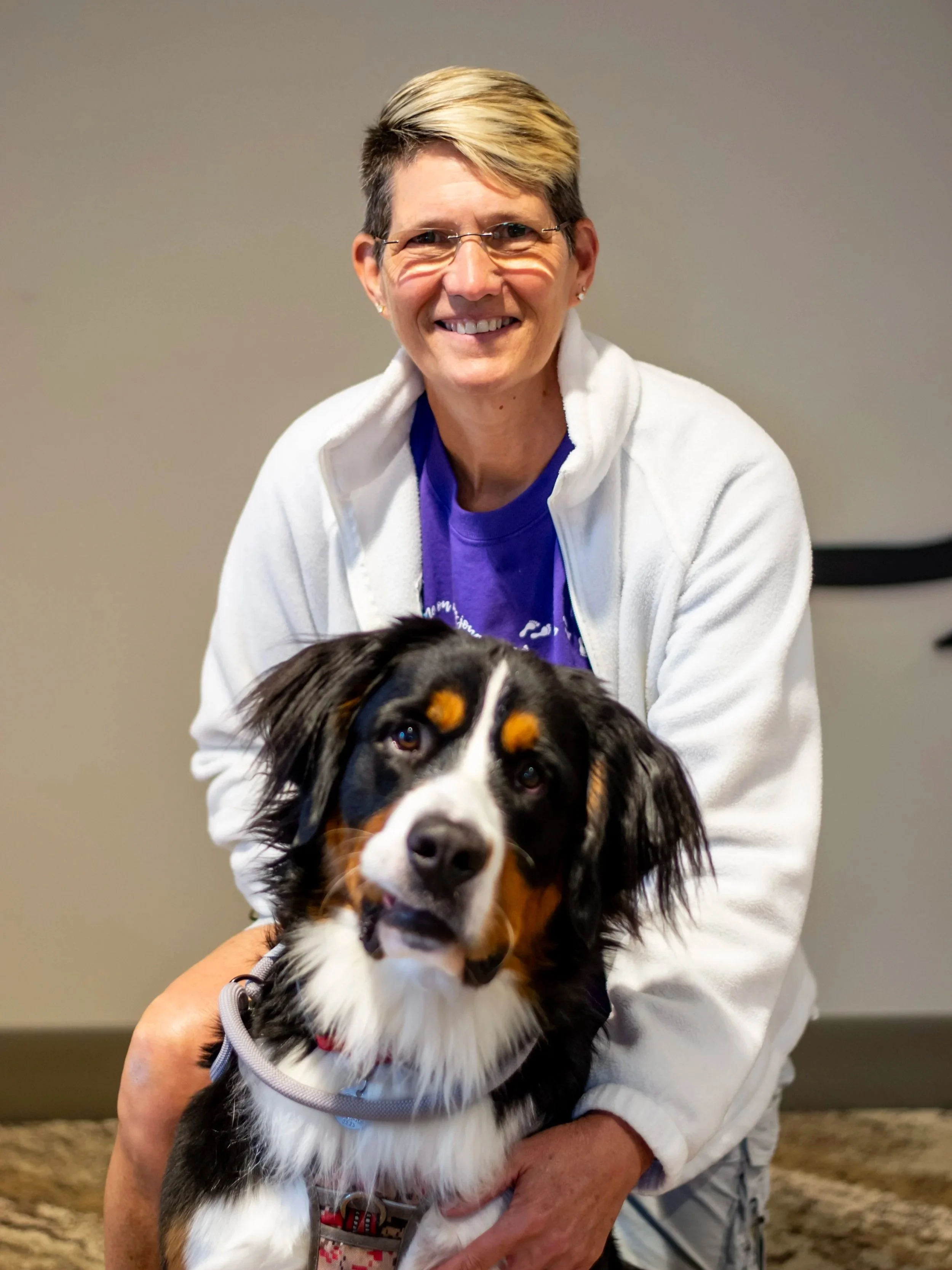 A woman with short hair and glasses smiling, holding an Australian Shepherd puppy with black, white, and tan fur, in an indoor setting with a plain wall background.