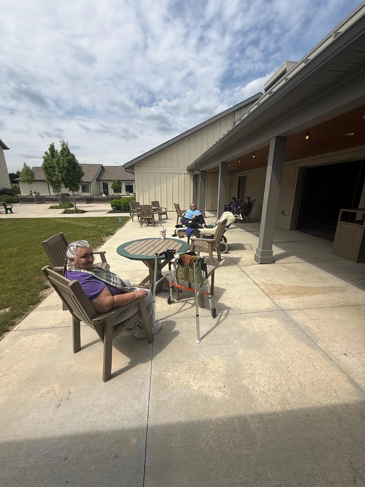 People sitting and lying on a covered outdoor patio with chairs and tables, in front of a beige building under a partly cloudy sky.
