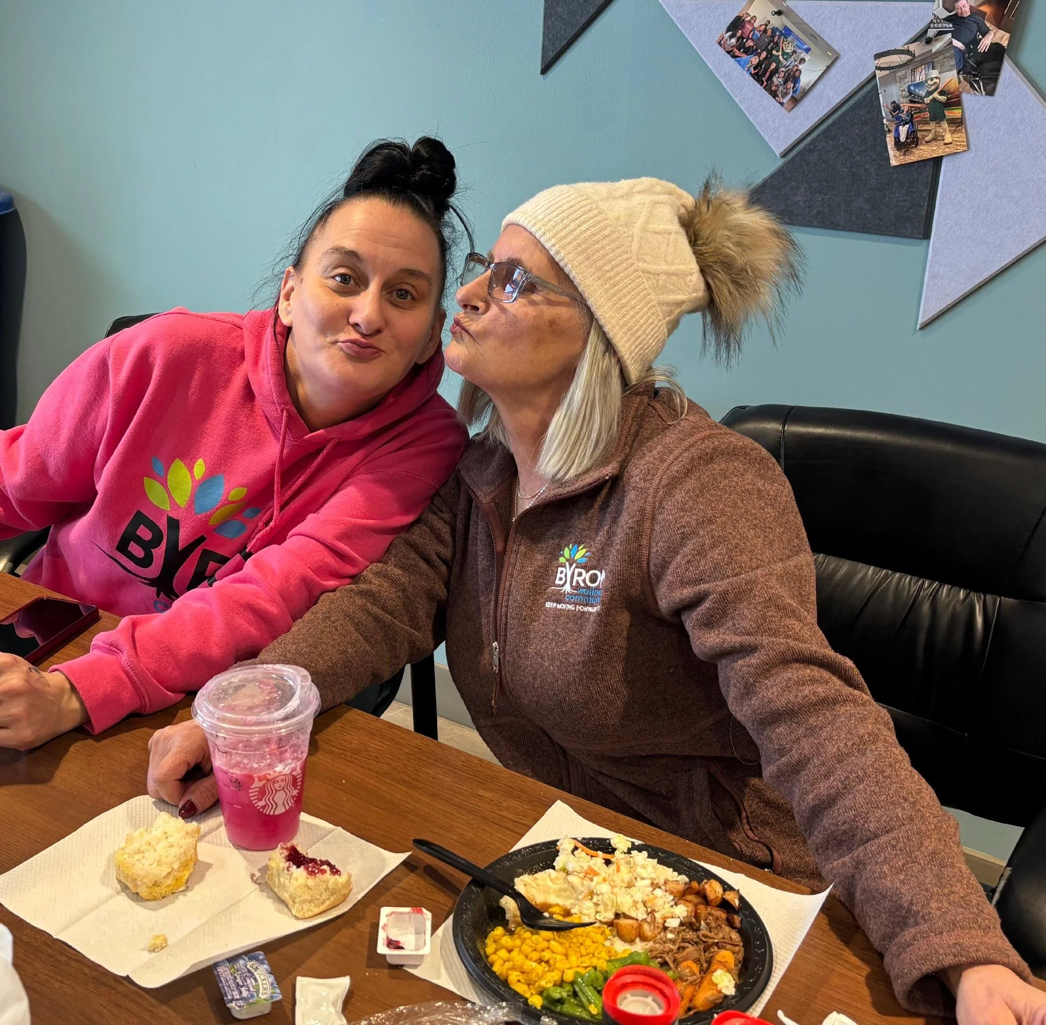 Two women sitting at a table with plates of food, cupcakes, and drinks, smiling and leaning towards each other for a photo, in a room with a teal wall and photos pinned on a bulletin board in the background.