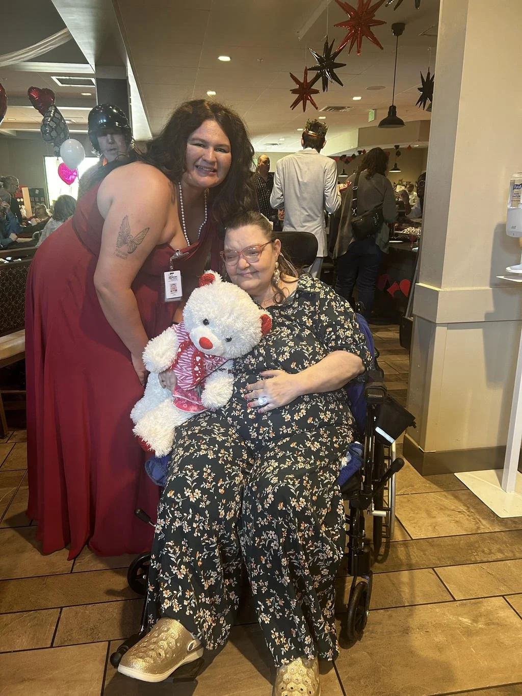 A woman in a red dress standing next to a woman in a wheelchair holding a teddy bear, in a decorated restaurant or cafe with other people in the background.