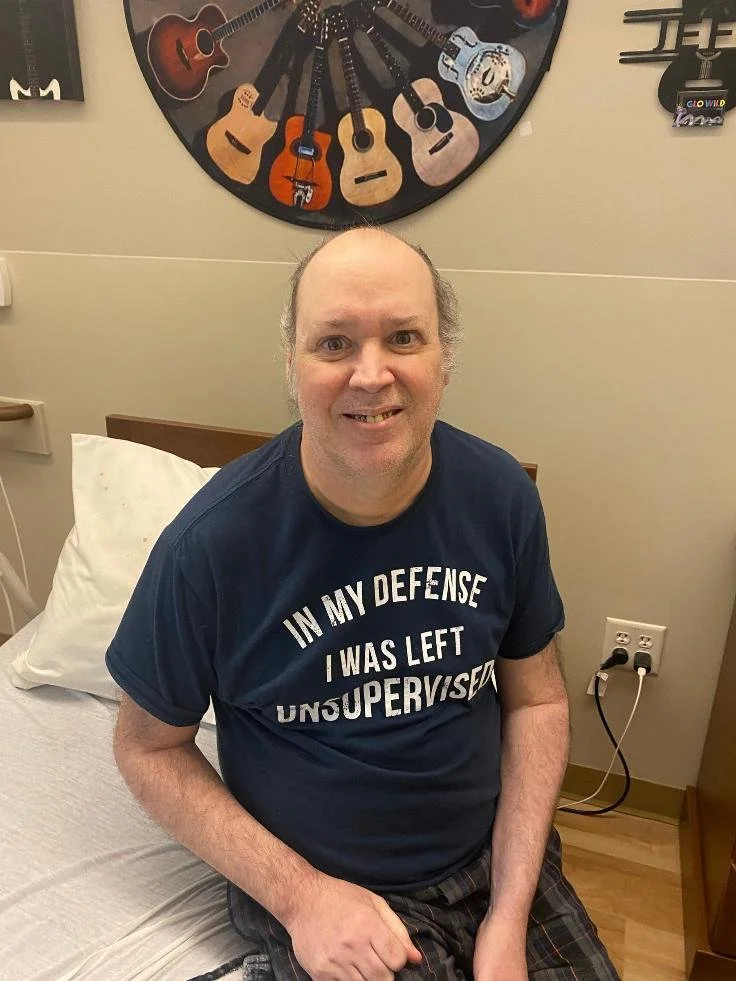 A man sitting on a hospital bed in a room with a wall-mounted display of various guitars in the background. He is wearing a navy T-shirt with white text that reads 'IN MY DEFENSE I WAS LEFT UNSUPERVISED'.
