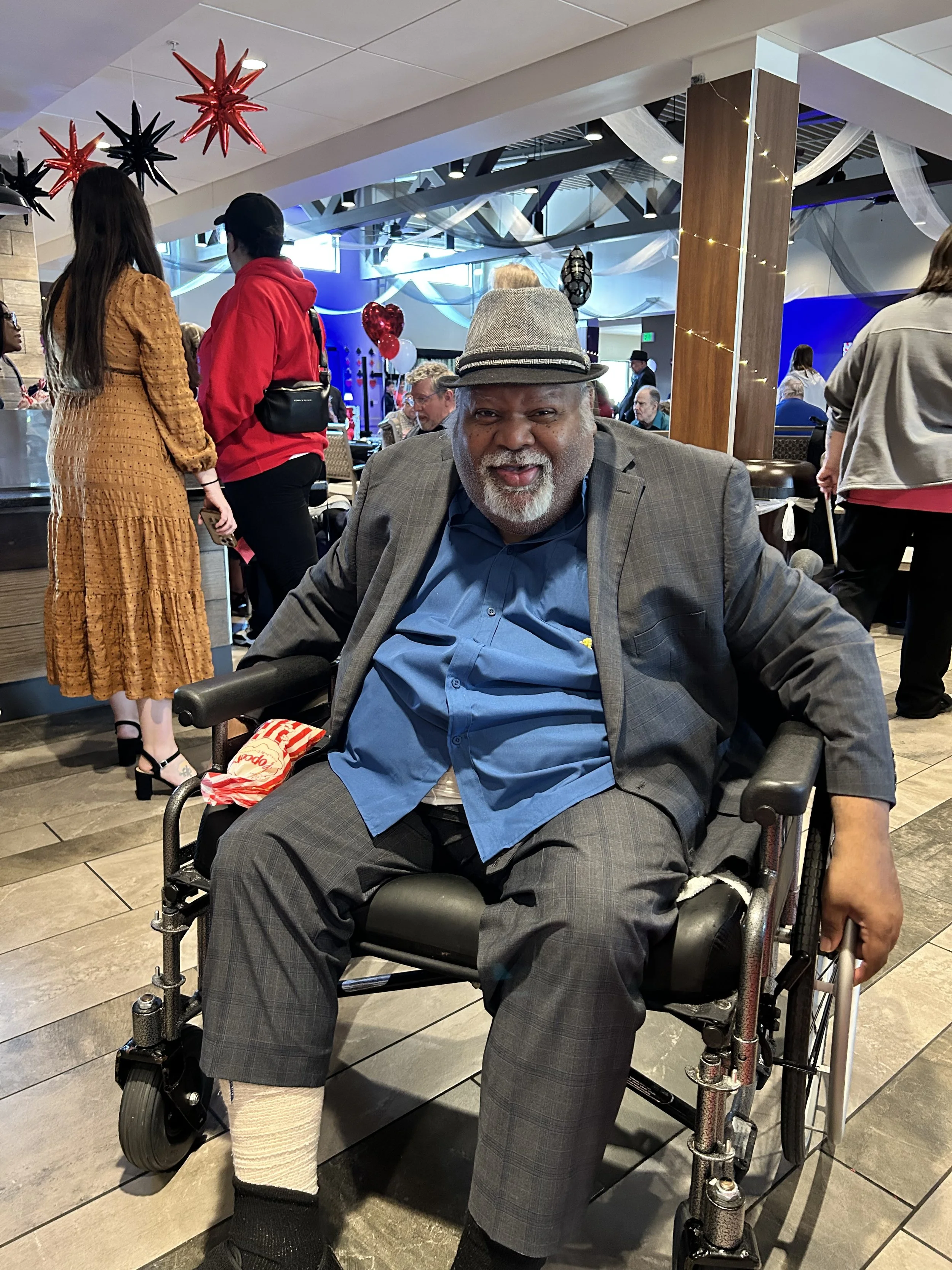 An elderly man with gray hair and beard in a wheelchair at a decorated indoor event with festive decorations, balloons, and people in the background.