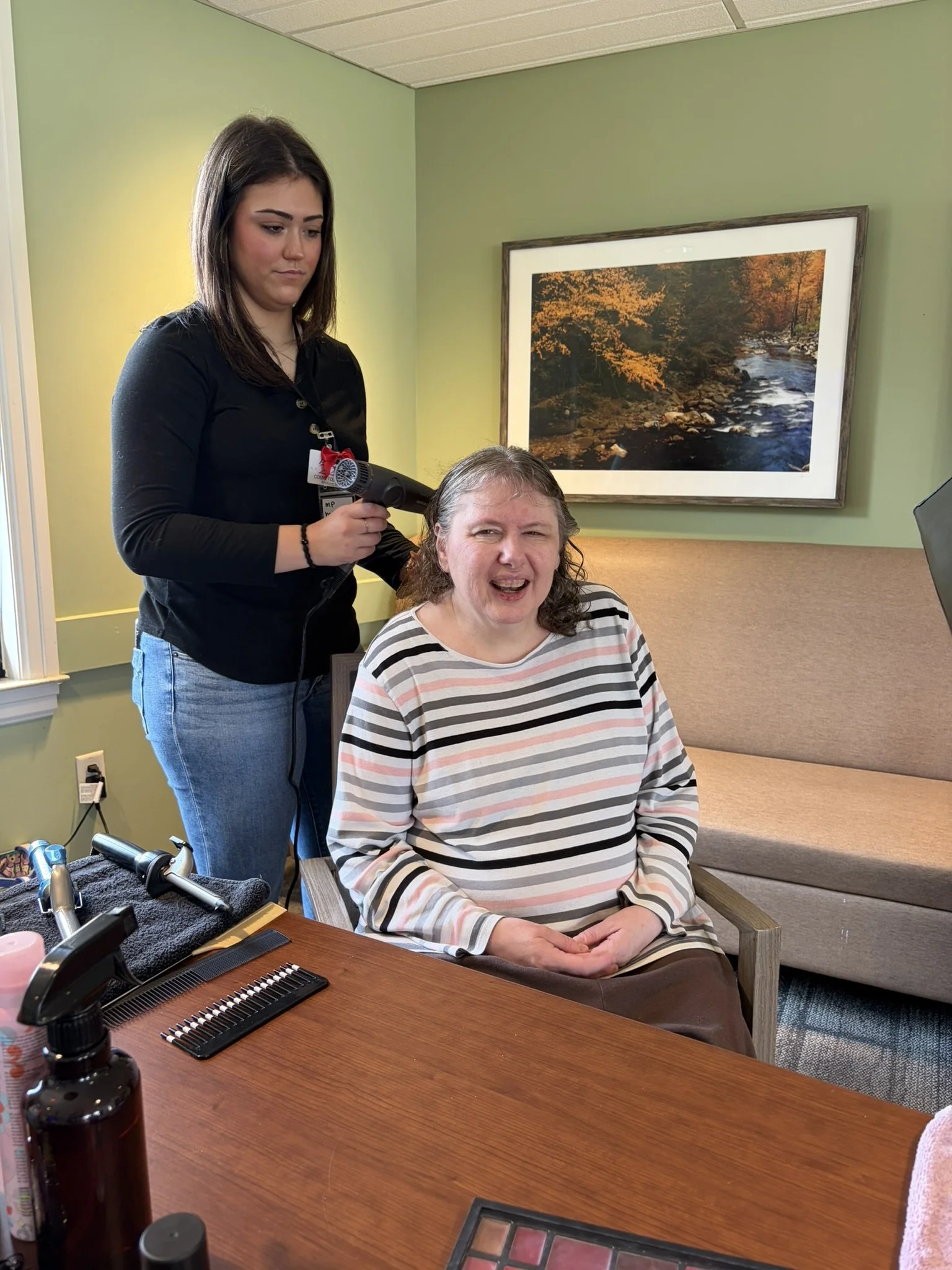 A woman sitting in a chair getting her hair styled with a hairdryer by a stylist in a salon or healthcare setting, with a large framed autumn landscape painting on the wall behind them.