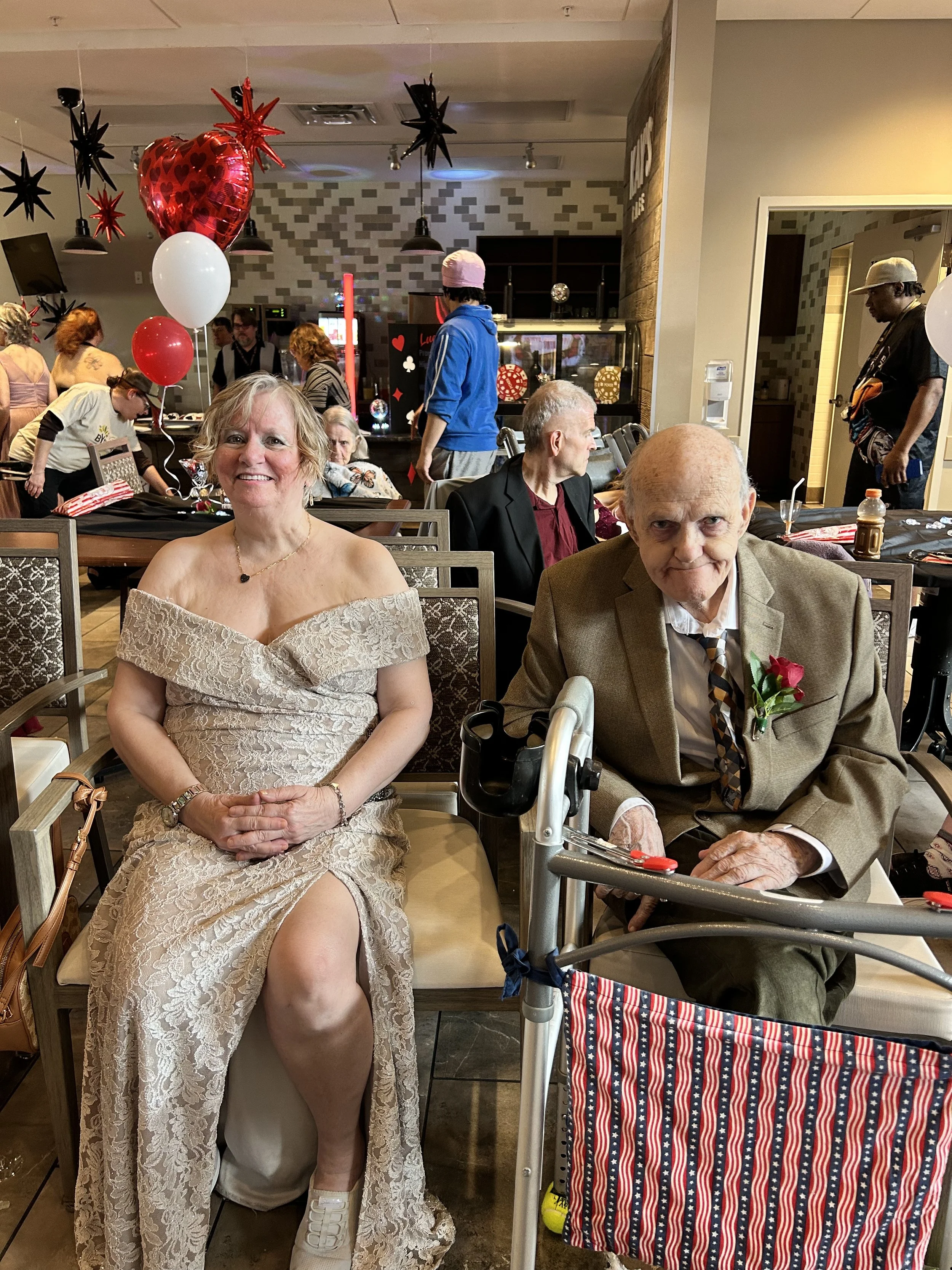 A woman in a lace dress and a man in a tan blazer and tie sitting at a table during a celebration with balloons and decorations in the background.