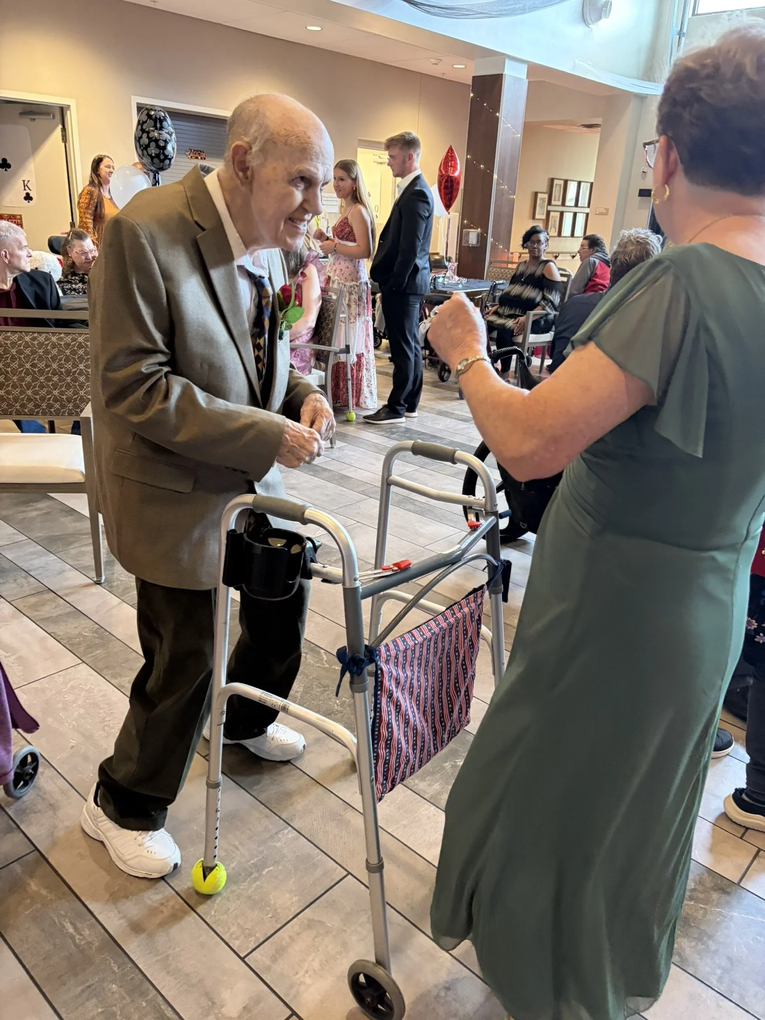 An elderly man with a walker playing tennis with a yellow ball in a community indoor setting. He is dressed in a beige blazer, white shirt, and dark pants. Several people are sitting and standing around, and some are wearing formal attire. Bright bal