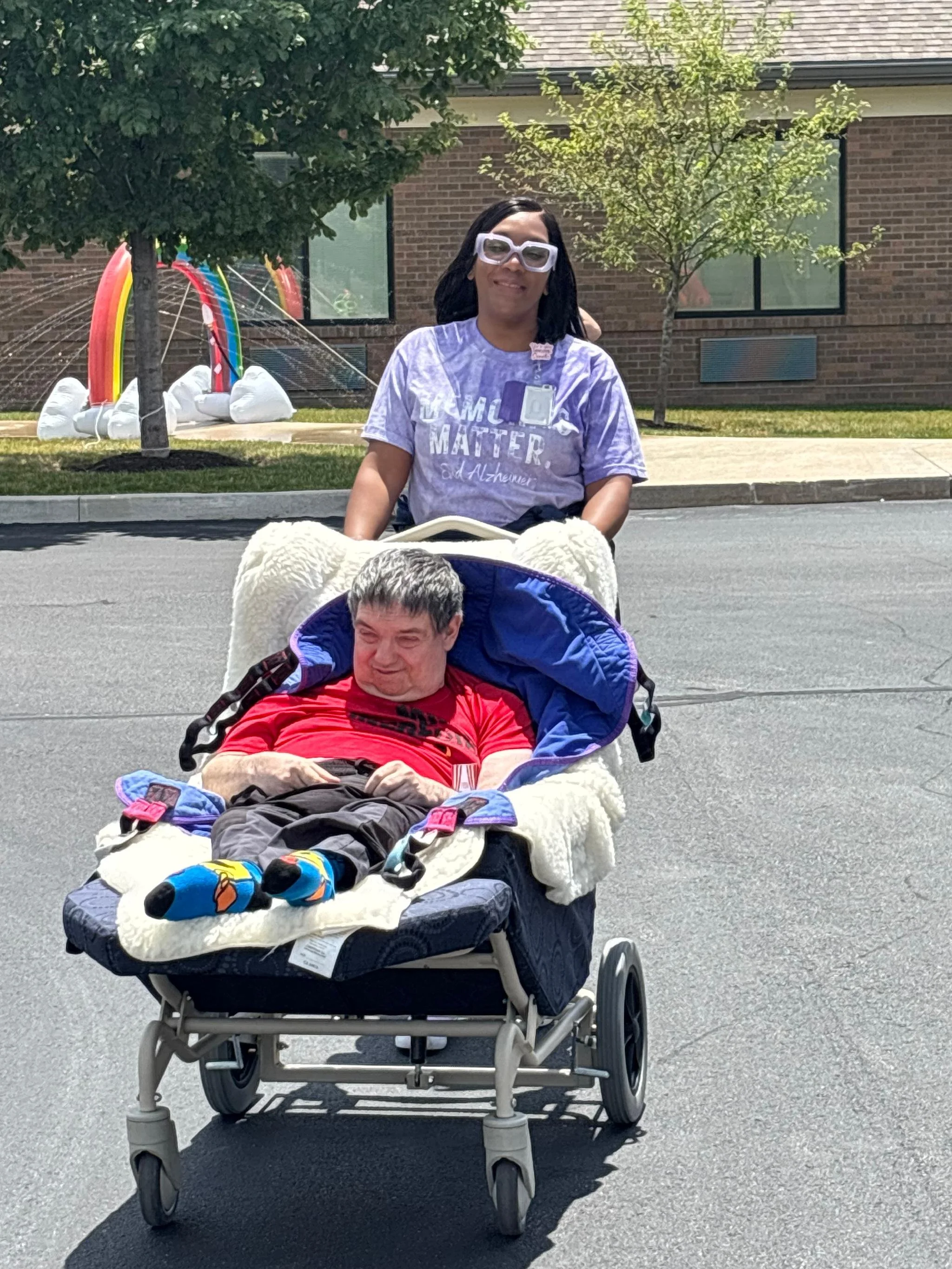 A woman in a wheelchair wearing glasses and a purple shirt pushes a man lying in a decorated wagon on a street. The wagon has stuffed animals and clothes, and the woman wears a shirt that supports Alzheimer's awareness.