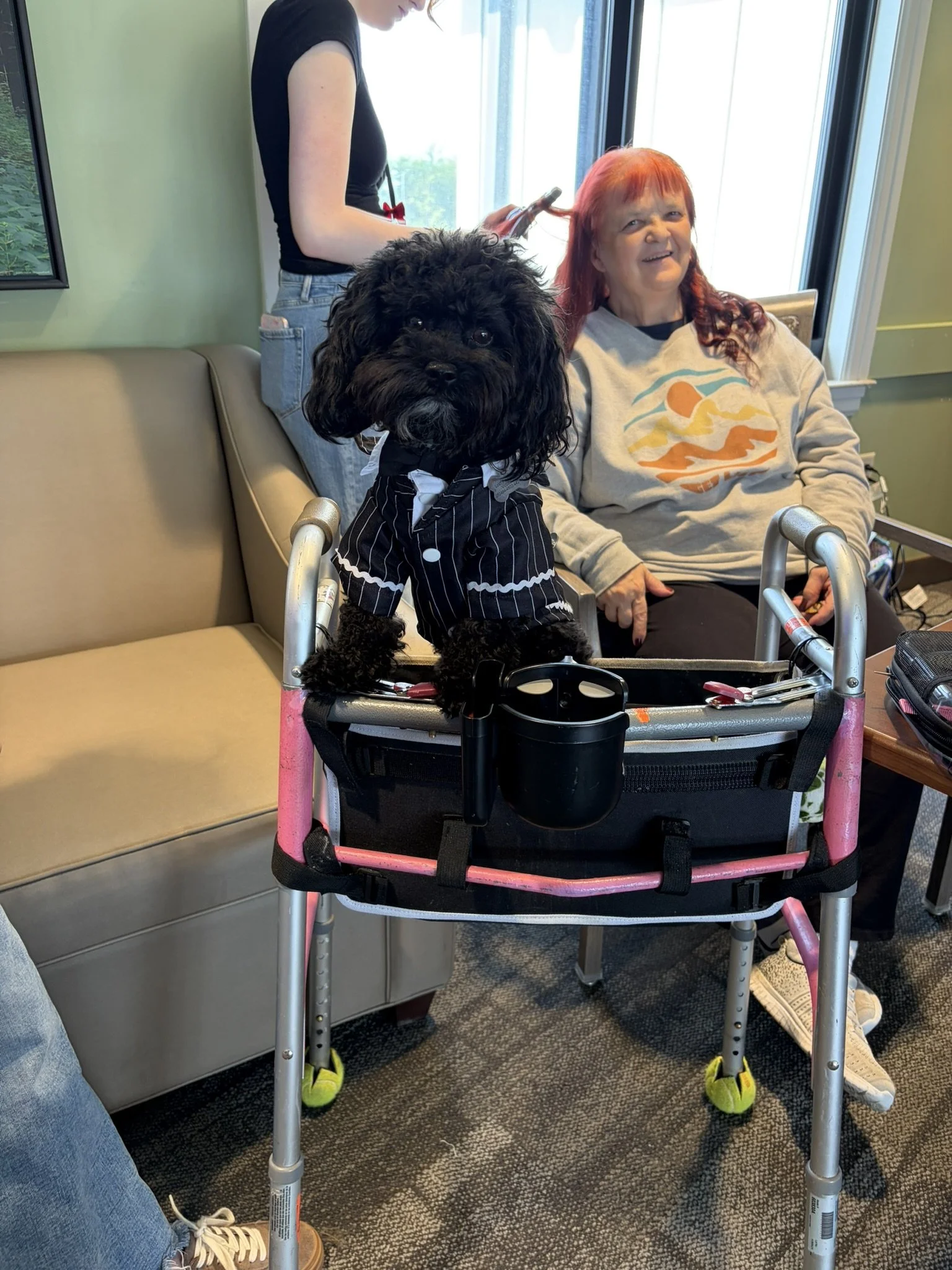 A woman with red hair sitting in a wheelchair at a hospital or care facility, smiling, as a nurse or caregiver with short hair and wearing a black shirt and jeans applies a hair treatment. There is a small black dog with curly fur wearing a black and