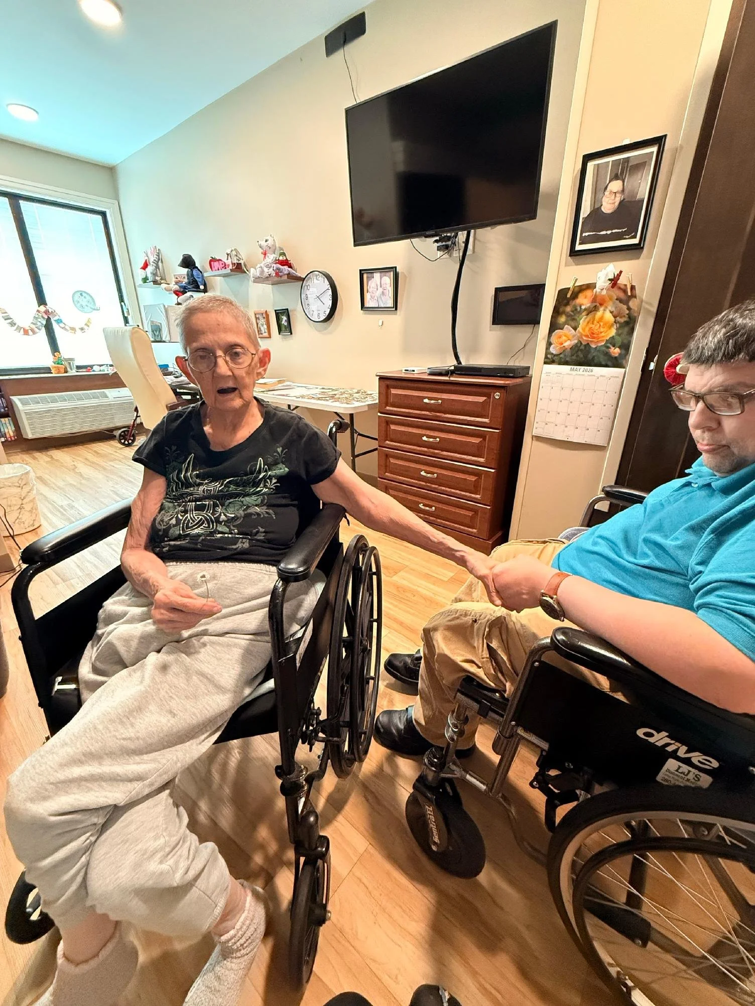 An elderly woman in a wheelchair and a middle-aged man in a wheelchair holding hands in a living room, with a TV, bookshelf, and decorated window in the background.