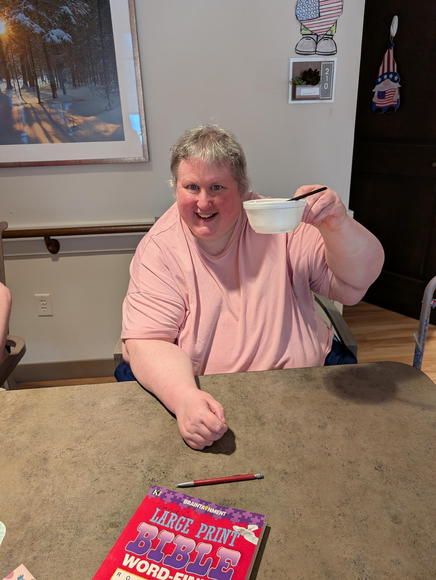 A person with short blonde hair, wearing a pink T-shirt, smiling and holding a white bowl and a black spoon. They are sitting at a table with a red and purple book titled 'Large Print Bubble Words' nearby. The background features framed artwork, wall