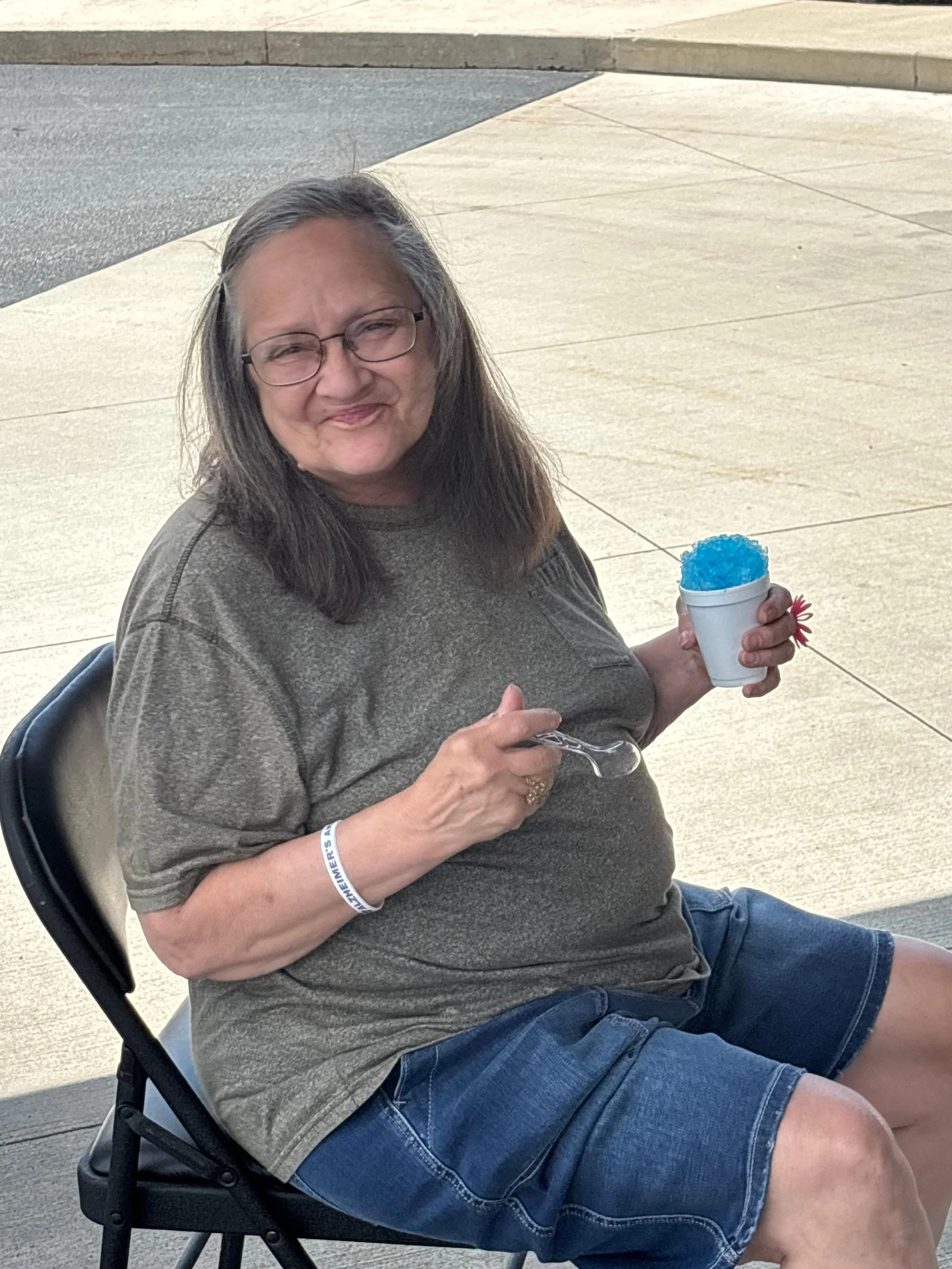 A woman with gray hair, glasses, wearing a gray shirt and denim shorts, sitting on a chair outdoors, holding a cup of blue snow cone with a pink straw, smiling at the camera.
